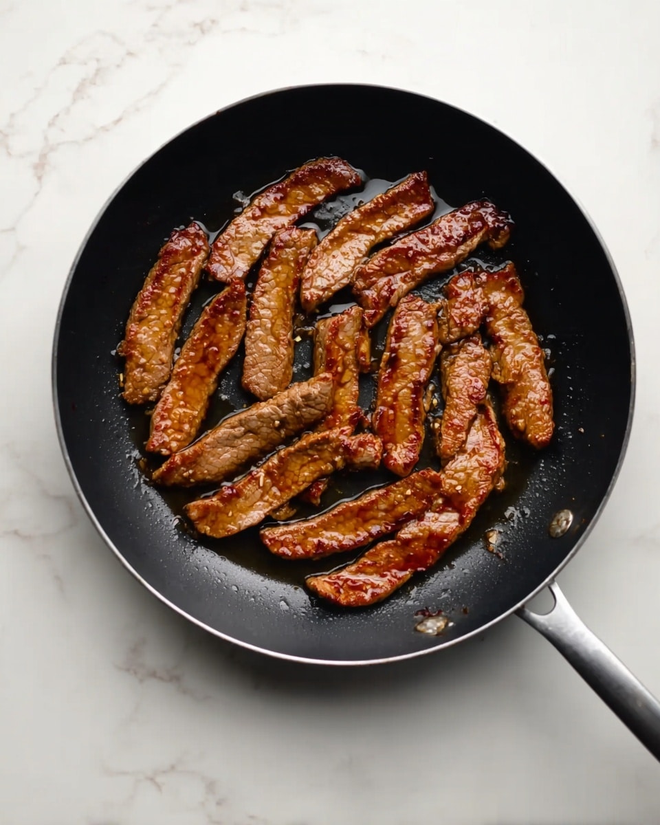 A black frying pan contains several strips of cooked meat with a brown color and a slightly shiny surface from the sauce or oil. The meat strips are arranged in a single layer, some overlapping slightly. The pan sits on a white marbled surface, and the handle extends outward, partially visible. The photo taken with an iphone --ar 4:5 --v 7