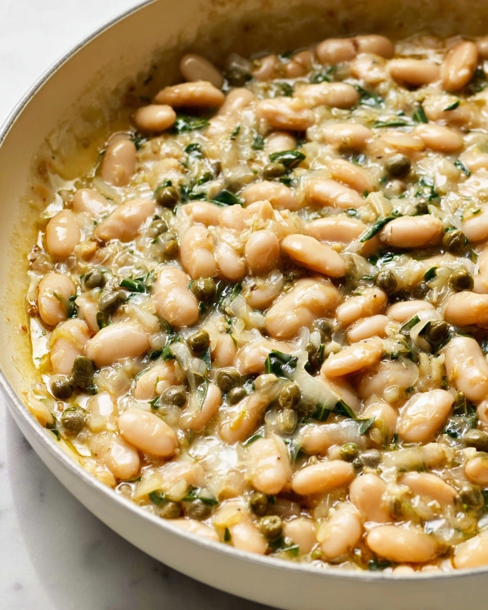 A close-up view of a creamy white bean dish in a white pan, showing one main layer made of soft, light beige beans mixed with small dark green capers and finely chopped white onions, all coated in a thick, glossy sauce that looks smooth and slightly oily, sitting on a white marbled surface photo taken with an iphone --ar 4:5 --v 7