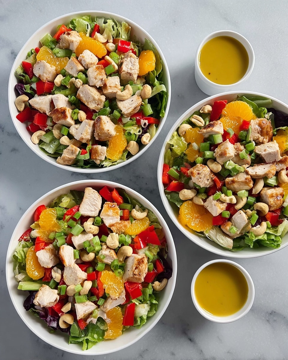 Three white bowls are filled with a colorful salad placed on a white marbled surface. Each bowl shows a base layer of mixed greens with bright orange mandarin slices and chopped red bell peppers spread across. On top of this are chunks of light brown grilled chicken pieces scattered evenly. The salad is garnished with chopped green onions and light beige cashew nuts, adding texture and detail. To the right of the bowls, three small white cups hold a yellow salad dressing. The photo taken with an iphone --ar 4:5 --v 7