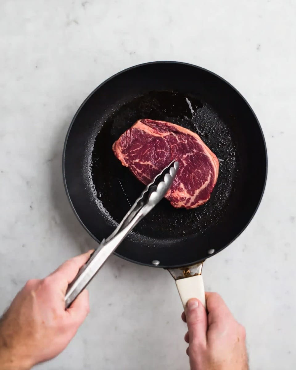 A close-up image shows a woman's hand holding metal tongs to place a thick piece of raw marbled steak in a black frying pan. The pan is held by another hand gripping the white handle. The background is a white marbled surface. The steak has deep red and white marbled textures, and the pan has slight oil reflections on its black surface. Photo taken with an iphone --ar 4:5 --v 7
