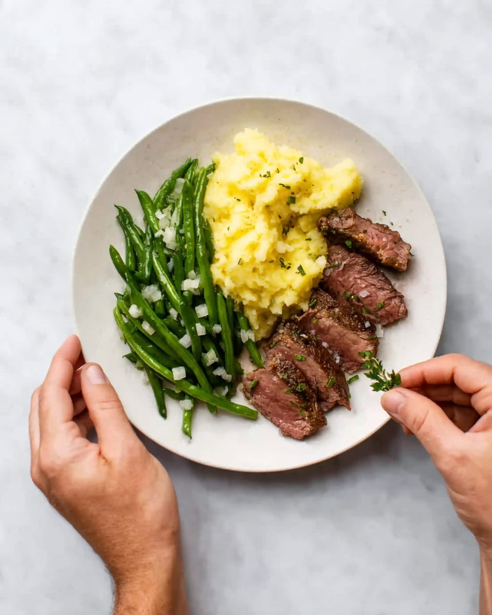 A white plate holds three main layers of food arranged side by side: on the left, bright green beans with small white bits scattered on top, in the middle, a serving of soft yellow mashed potatoes with a slightly uneven texture, and on the right, slices of brown steak arranged in a neat row with a hint of pink inside and small green herbs sprinkled on top. The plate is held by a woman's left hand at the bottom, while a man's right hand is placing a tiny green herb onto the steak. The background is a white marbled surface. photo taken with an iphone --ar 4:5 --v 7