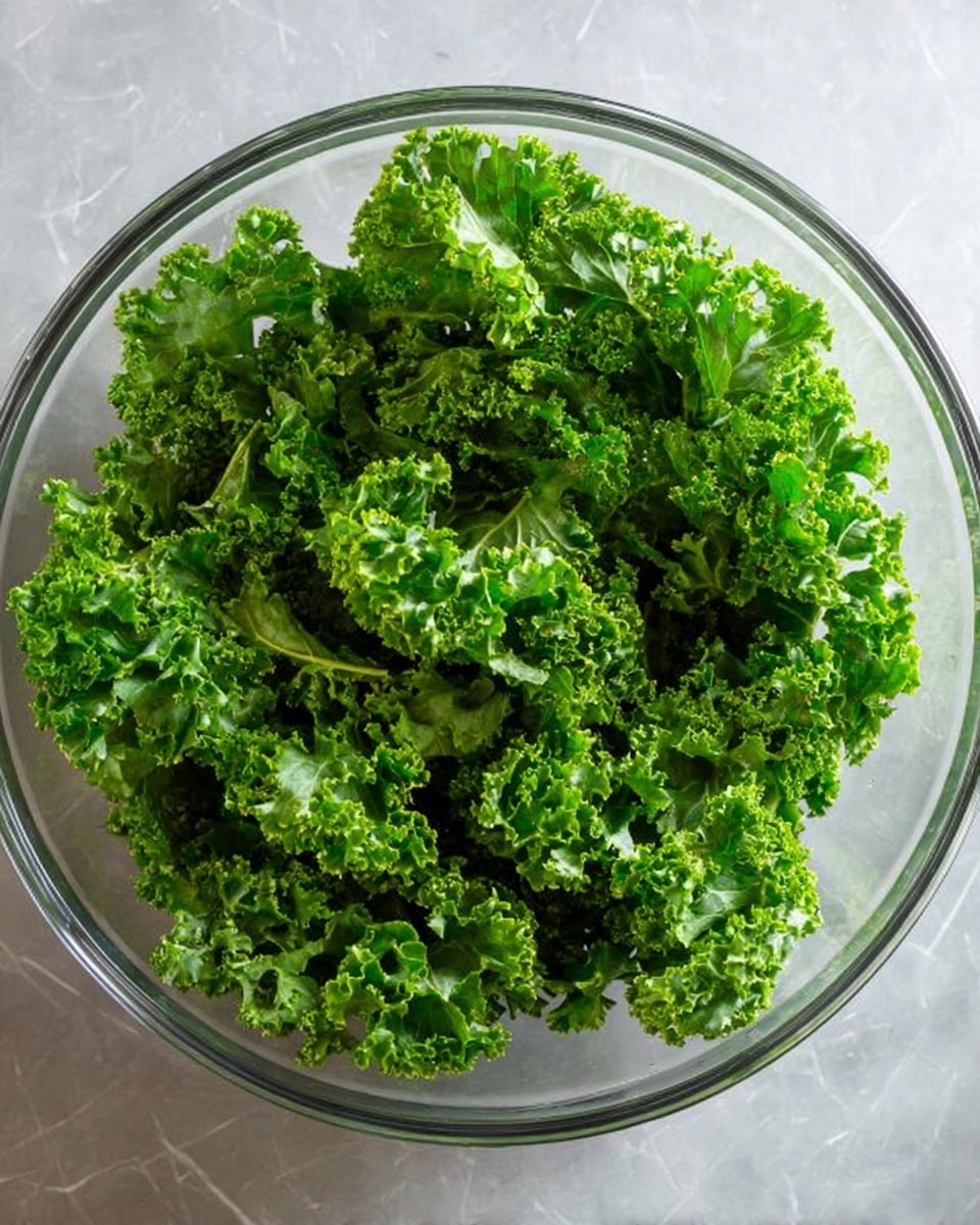 A clear glass bowl is filled with a single layer of fresh green kale leaves, showing their curly texture and bright green color. The bowl sits on a white marbled surface, and the kale covers the entire inside with no other ingredients visible. The kale looks fresh and ready to use. photo taken with an iphone --ar 4:5 --v 7