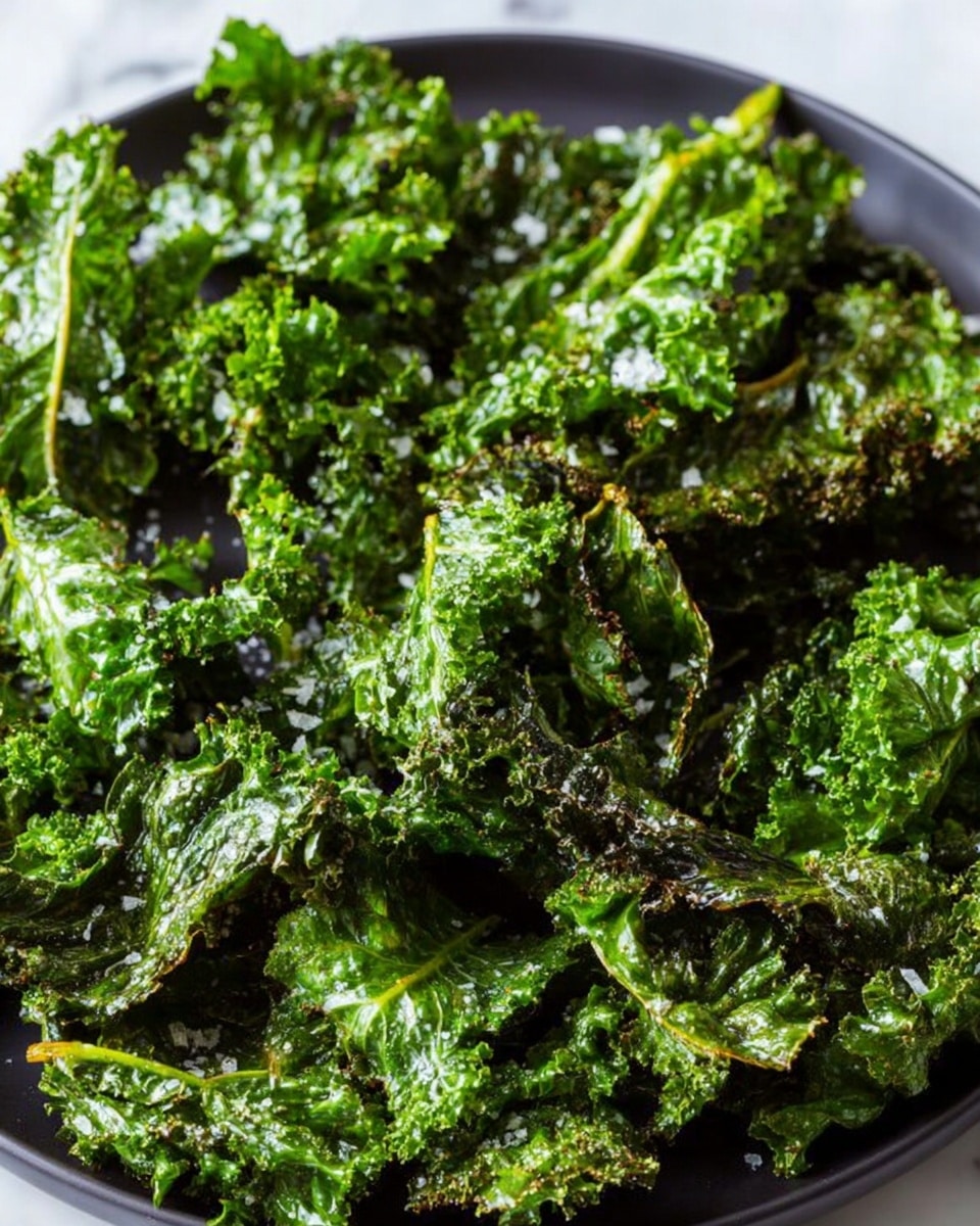 A close-up view of a white plate filled with crispy kale chips showing multiple layers of bright green, wrinkled and curly kale leaves with some darker toasted edges, and small white sea salt flakes sprinkled on top. The texture looks crunchy and slightly glossy with light and shadow creating depth among the kale leaves. The background is a white marbled surface. photo taken with an iphone --ar 4:5 --v 7