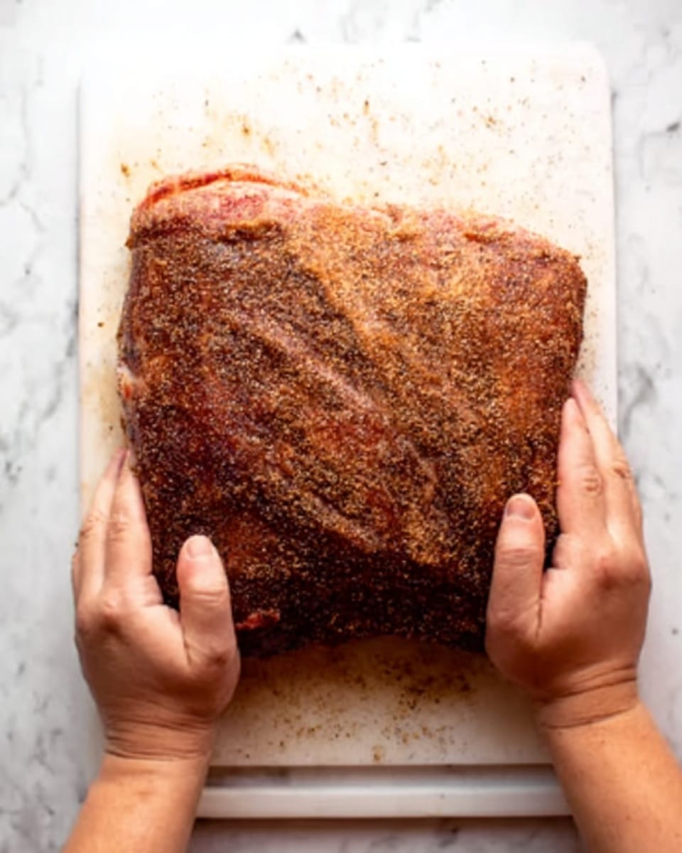 A large slab of seasoned meat with a dark brown, slightly rough textured outer layer is being held at the top edges by a pair of hands, visible from above. The meat sits on a white cutting board placed on a white marbled surface. The seasoning on the meat surface shows a mix of spices giving it a coarse, grainy appearance. Photo taken with an iphone --ar 4:5 --v 7