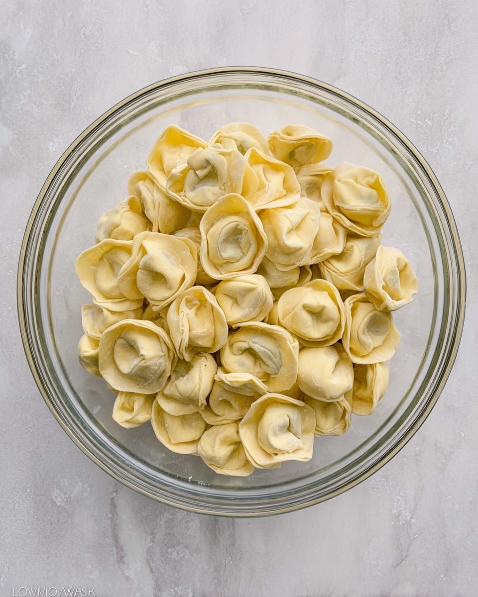 A clear glass bowl filled with many pieces of uncooked tortellini pasta, each having a pale yellow color with smooth, slightly folded edges. The pasta pieces are layered randomly inside the bowl, showing slight uneven shapes and textures typical of fresh pasta. The bowl sits on a white marbled surface with subtle gray veins and an overall clean look. The photo is taken from above, capturing the round shape of the bowl and the delicate details of each tortellini piece photo taken with an iphone --ar 4:5 --v 7