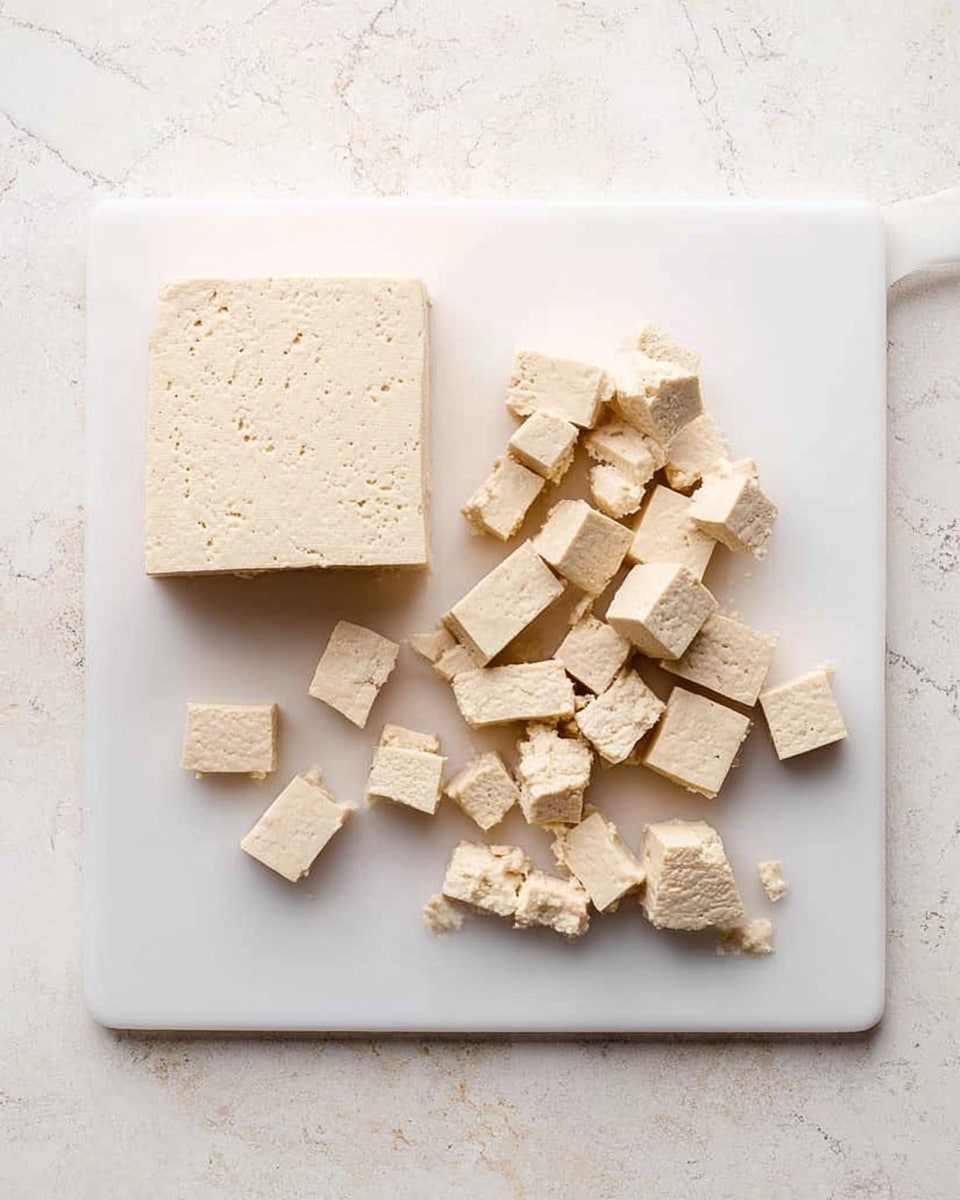 The image shows a white cutting board on a white marbled surface. On the board, there is a block of light beige tofu that is partially cut into small cubes. The whole block is on the left side, and scattered pieces of tofu cubes are arranged on the right side in a loose, uneven pattern. The texture of the tofu is smooth with small holes, typical for tofu. The lighting is bright and natural, highlighting the pale color and soft texture of the tofu. photo taken with an iphone --ar 4:5 --v 7