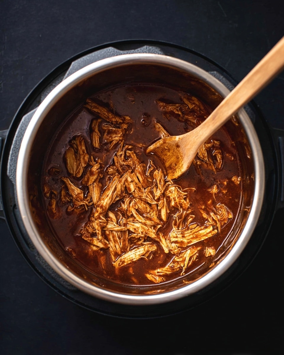 An overhead view of shredded cooked chicken soaking in a deep, rich brown sauce inside a silver metal pot. A wooden spoon with a smooth texture is scooping some of the chicken, showing strands of tender meat mixed with bits of sauce. The pot sits on a black surface. photo taken with an iphone --ar 4:5 --v 7