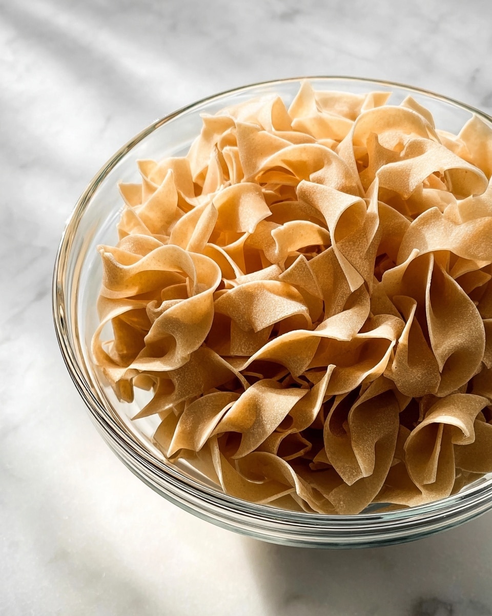 A clear glass bowl filled with dry egg noodle pasta, showing many layers of light brown, wavy, and curly noodle ribbons stacked closely together. The bowl sits on a white marbled texture surface, with a soft natural light highlighting the smooth, slightly curved edges of the noodles and casting gentle shadows inside the bowl. photo taken with an iphone --ar 4:5 --v 7