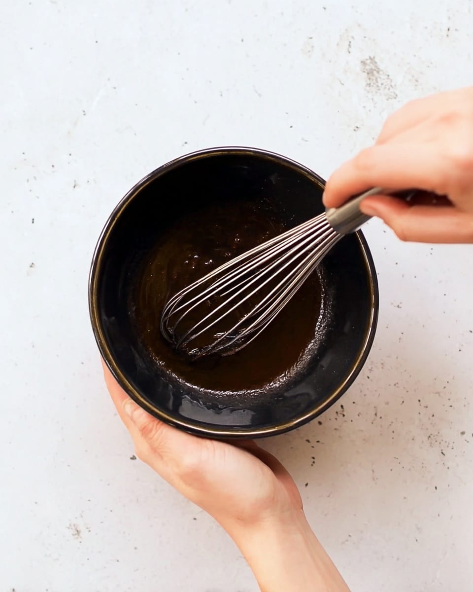 A woman's hand holds a black bowl filled with a dark brown mixture that looks smooth and shiny. Inside the bowl, another woman's hand is mixing the contents with a silver whisk that has thin metal wires. The background shows a clean white marbled surface, creating a simple and bright setting. Photo taken with an iphone --ar 4:5 --v 7