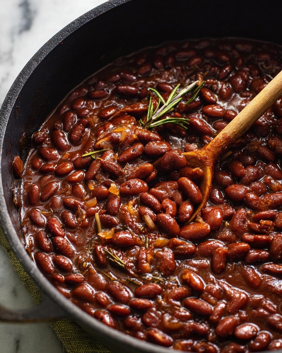 A close-up view of a black pan filled with dark brown cooked beans in a rich, thick sauce with visible small chopped onions and sprigs of rosemary mixed in. A wooden spoon rests inside the pan, partly submerged in the beans, showing a glossy texture and a deep reddish-brown color with bits of onion and herbs spread evenly throughout. The background shows a white marbled texture. Photo taken with an iphone --ar 4:5 --v 7
