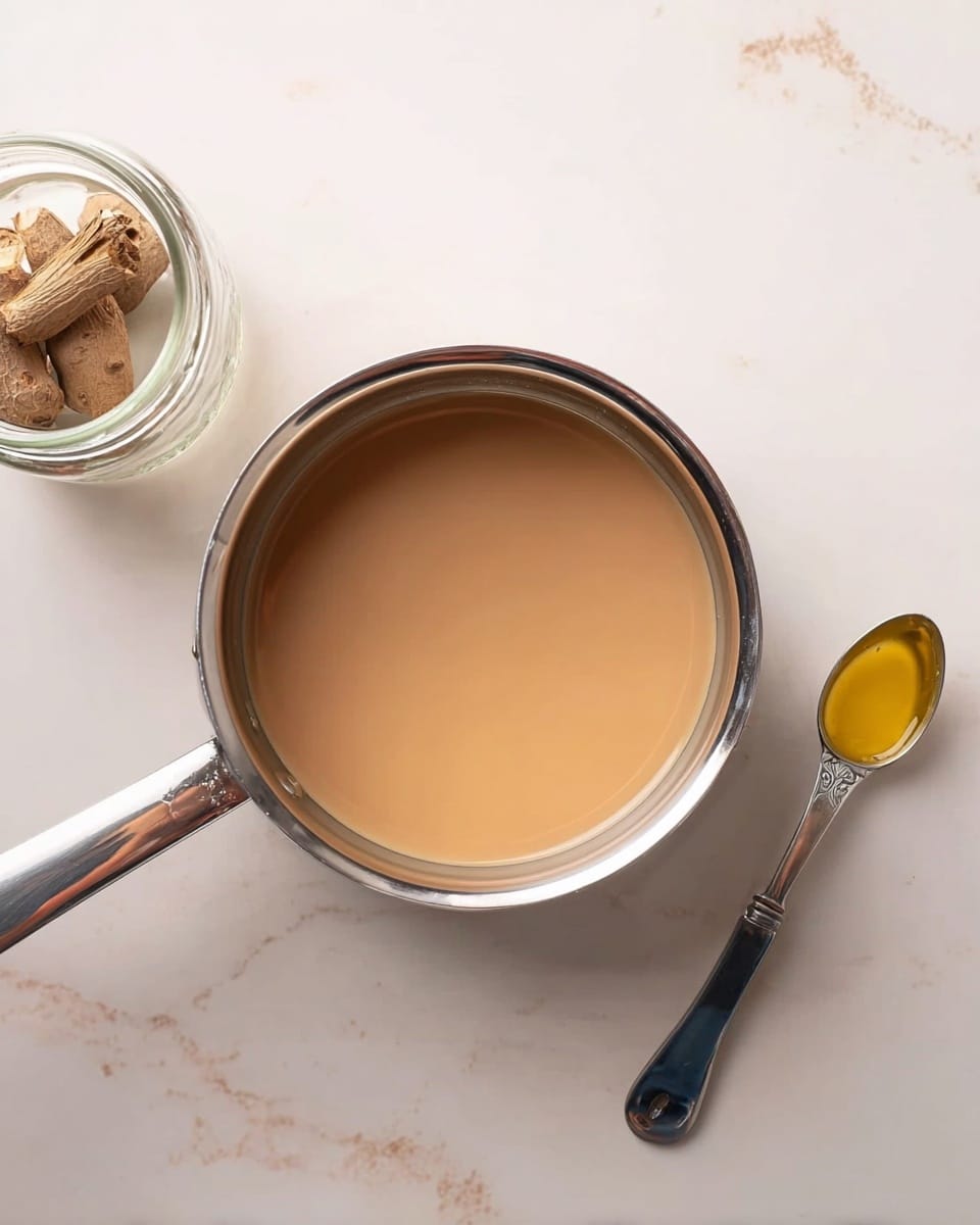 A small silver pan filled with smooth, light brown liquid, centered on a white marbled surface. To the left of the pan is a small clear glass jar with a few pieces of brown dried roots inside. To the right of the pan is a silver spoon with black grip holding a small amount of yellow liquid. The pan's handle extends to the right side of the frame. The scene is softly lit, highlighting the shiny metal of the pan and the smooth texture of the liquid photo taken with an iphone --ar 4:5 --v 7