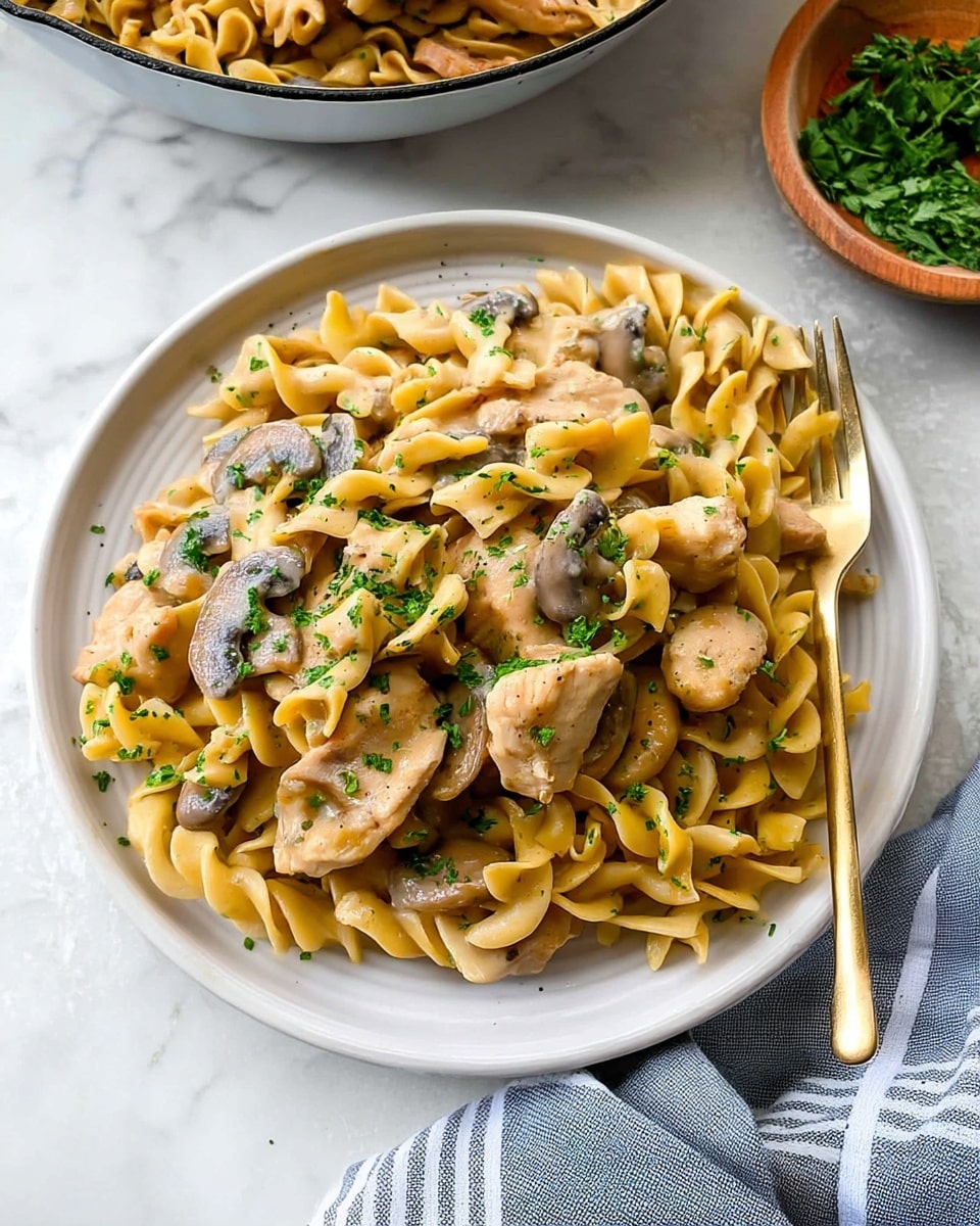 A white plate holds a serving of creamy pasta with three visible layers: the bottom layer is light yellow twisted egg noodles, the middle layer has light brown cooked mushroom slices scattered throughout, and the top layer features light tan sauce-coated pieces of chicken mixed with the noodles and mushrooms, all sprinkled with small green parsley bits. A gold fork rests on the right side of the plate, and the whole scene is set on a white marbled surface with a gray and white striped cloth nearby. In the background, part of a pan with more creamy pasta and a small wooden bowl of chopped parsley can be seen. photo taken with an iphone --ar 4:5 --v 7