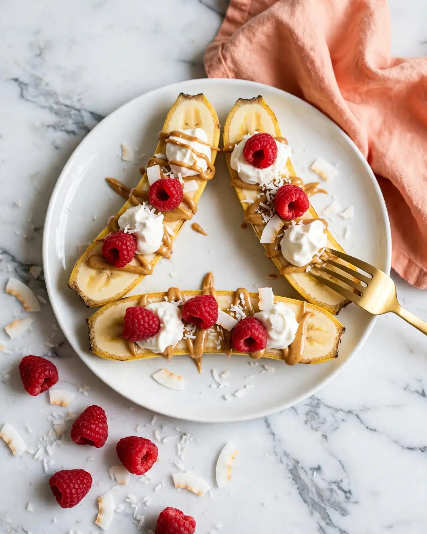 The image shows a white plate on a white marbled surface, holding three banana halves that are sliced lengthwise and arranged in a triangle shape. The bananas are topped with a drizzle of light brown peanut butter, dollops of white whipped cream, and scattered coconut flakes. Bright red raspberries sit on top of the whipped cream and bananas, adding vibrant color. Some raspberries and coconut flakes spill onto the white marbled surface around the plate. A gold fork rests on the right side of the plate, touching one banana, with a peach-colored cloth napkin underneath the plate toward the top right. The photo is taken with an iphone --ar 4:5 --v 7