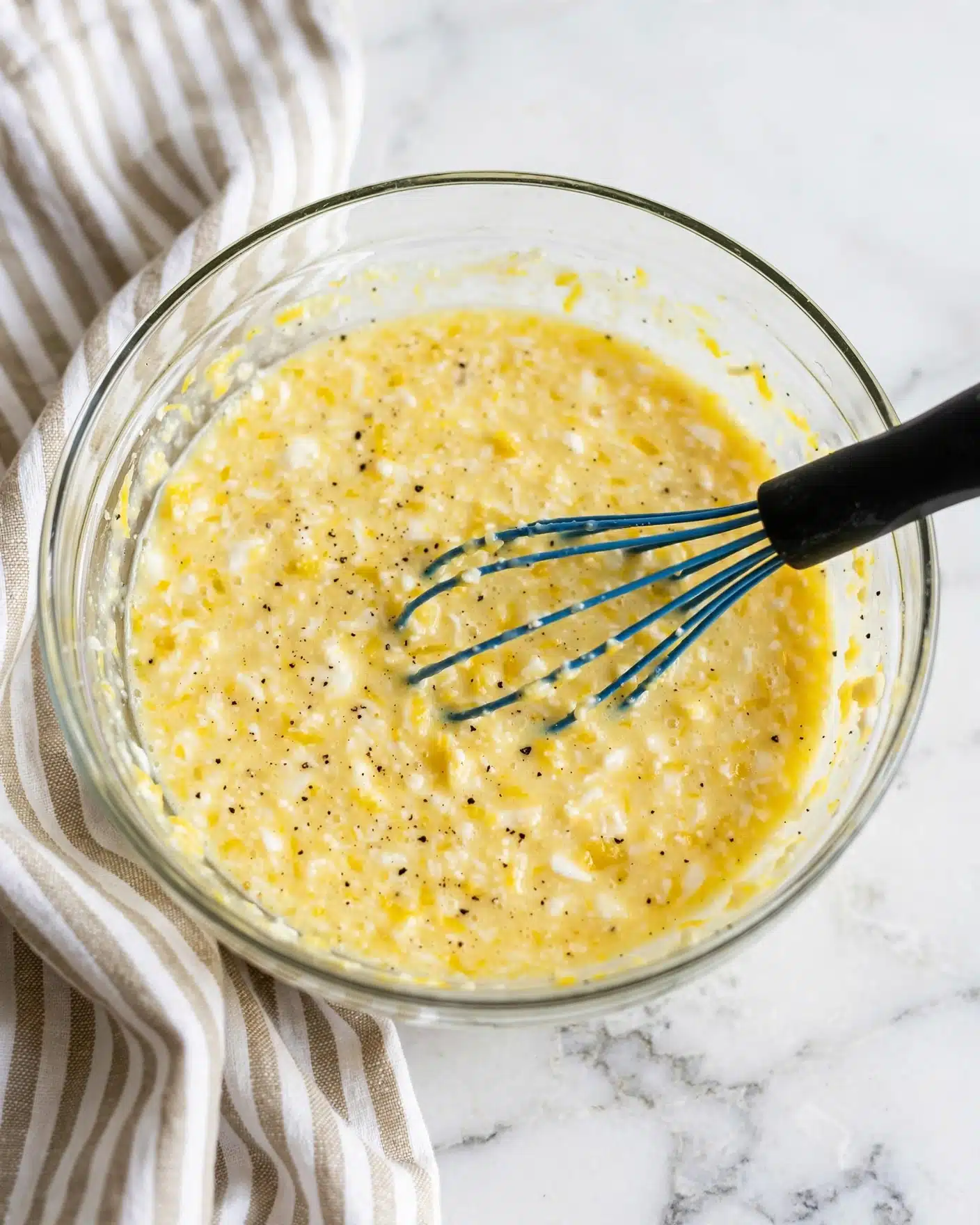A clear glass bowl holds a mixture of light yellow liquid with small bits of white and darker yellow, showing a slightly thick texture with some specks of black pepper. A blue whisk with a black handle is partly submerged in the mixture, resting inside the bowl near the center right. The bowl sits on a white marbled surface, with a soft, beige-and-white striped cloth partially under the bowl on the left side. The lighting is bright and natural, highlighting the smooth texture and colors of the mixture. Photo taken with an iphone --ar 4:5 --v 7