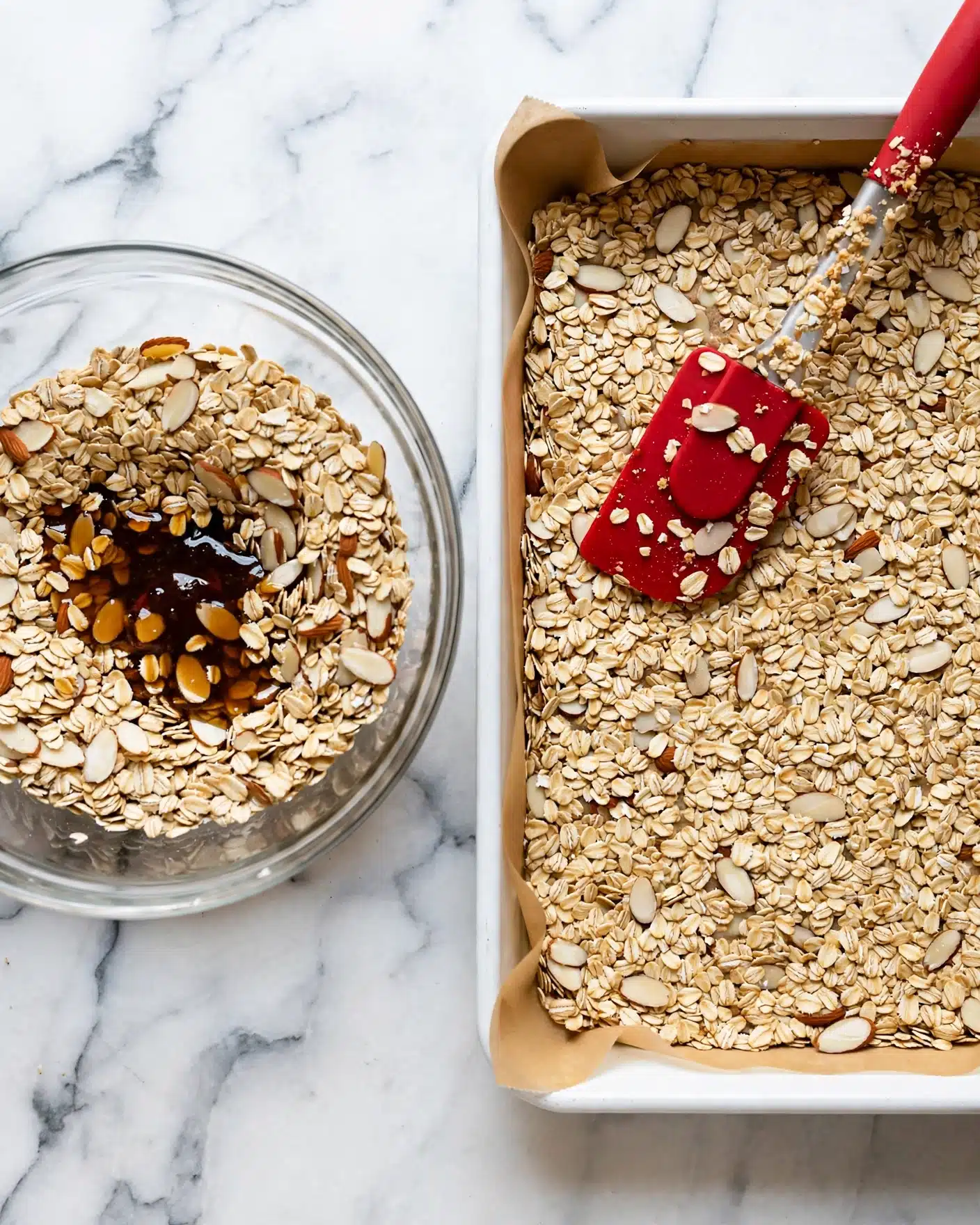The image shows two parts: on the left, a clear glass bowl filled with dry rolled oats mixed with sliced almonds and a dark syrupy liquid pooled in the middle; on the right, a white rectangular baking pan lined with light brown parchment paper, evenly filled and spread with the same oat and almond mixture, with a red rubber spatula resting on top, partially covered with oats. The background is a white marbled texture. photo taken with an iphone --ar 4:5 --v 7