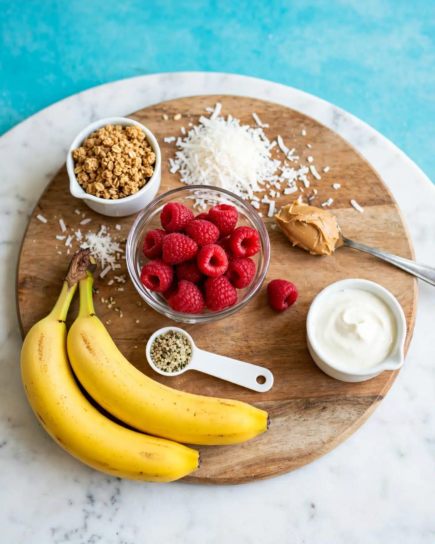 A round wooden board on a white marbled surface holds several ingredients arranged neatly: two bright yellow bananas are positioned at the bottom left, next to a white measuring spoon with small hemp seeds inside, resting on the board. A small white measuring cup filled with crumbly granola sits at the top left. Next to it is a small heap of shredded coconut, with some scattered around. In the center of the board is a clear glass bowl filled with fresh red raspberries, and one raspberry lies beside it. To the right of the bowl, a silver spoon with smooth peanut butter rests on the board, and below it, another white measuring cup holds a creamy white substance, likely yogurt or cream. The overall scene is clean, colorful, and well-organized, with a bright turquoise background visible beyond the board. photo taken with an iphone --ar 4:5 --v 7