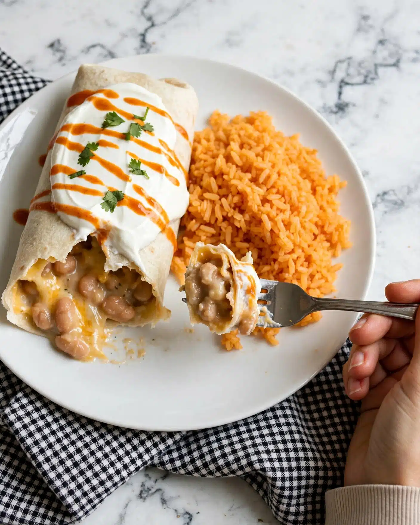 The image shows a white plate with one burrito partially opened on the left side, revealing creamy beans and melted yellow cheese inside the soft, light beige tortilla. On top of the burrito, there is a thick layer of white sour cream with thin orange sauce drizzled over it and small pieces of green cilantro leaves scattered for garnish. To the right of the burrito, there is a serving of fluffy orange rice with a slightly sticky texture. A silver fork is holding a bite-sized piece of the burrito in front of the rice, and part of a woman's hand is visible holding the fork. The plate is placed on a white marbled surface with a black and white checkered cloth partially under it. Photo taken with an iphone --ar 4:5 --v 7