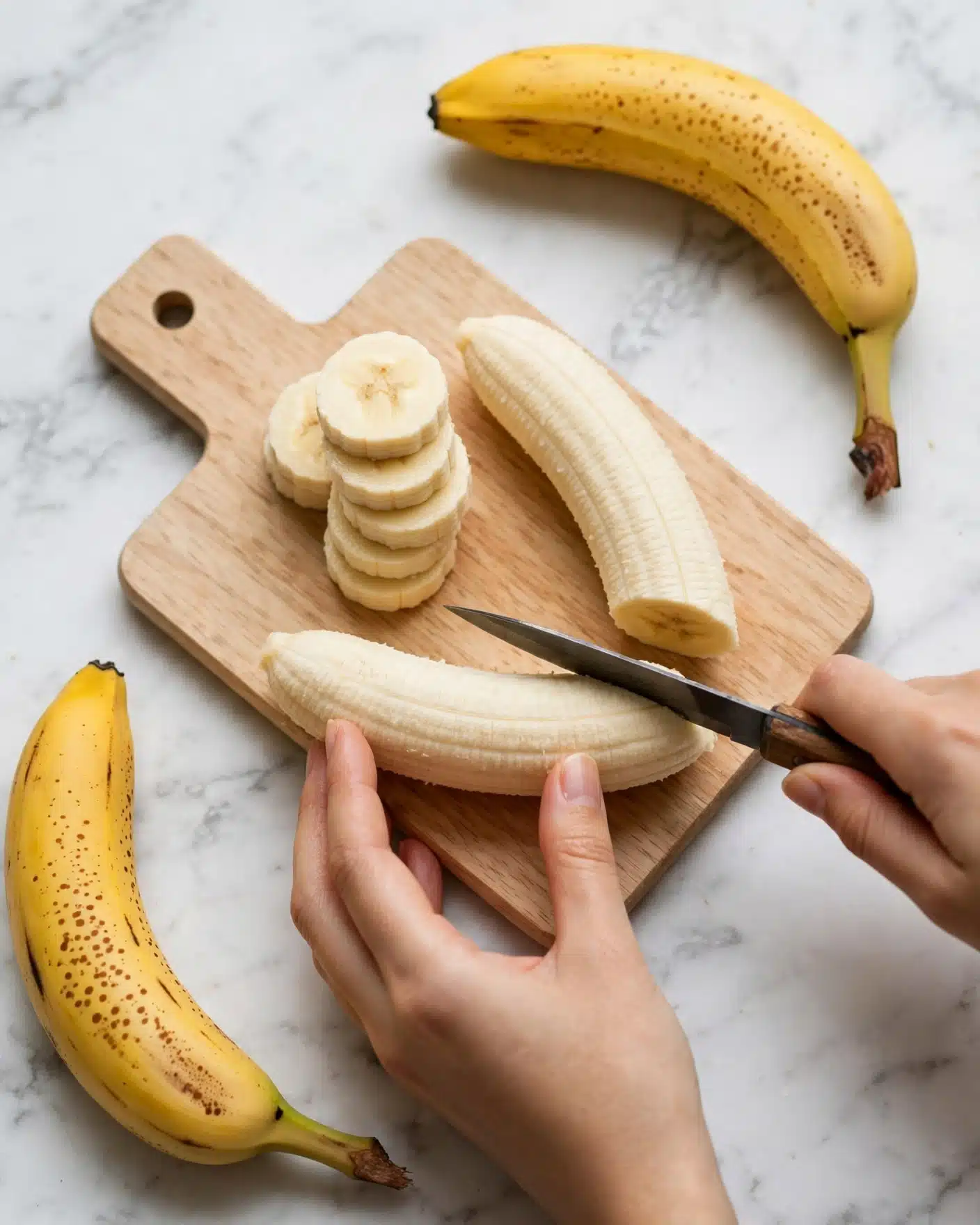 In the image, two peeled bananas are placed on a small light wooden cutting board with a round hole near the top left corner, lying on a white marbled surface. One banana is sliced into thick, even round pieces, stacked closely on the board showing a soft creamy inside, while the other is whole beside them. A woman's hand holds the banana gently at the bottom, while the other woman's hand grips a small black and wooden knife slicing through the banana. Two ripe speckled yellow bananas lay on the white marbled surface, one at the top right and one at the bottom left corner, creating balance in the frame. photo taken with an iphone --ar 4:5 --v 7