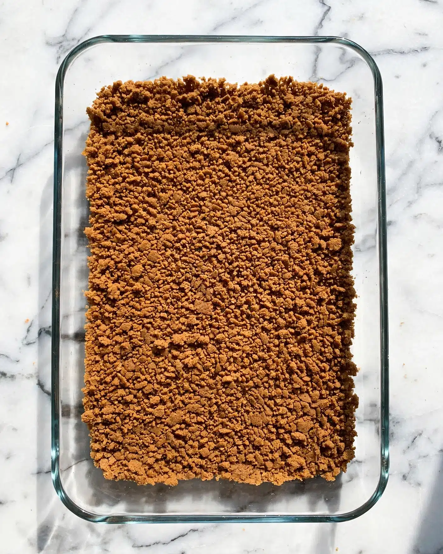 A clear glass rectangular baking dish is filled with one layer of finely crushed brown crumbs, evenly spread and pressed flat to cover the bottom of the dish. The crumbs have a rough yet compact texture, creating a solid base layer. The dish is placed on a surface with a white marbled texture. Photo taken with an iphone --ar 4:5 --v 7