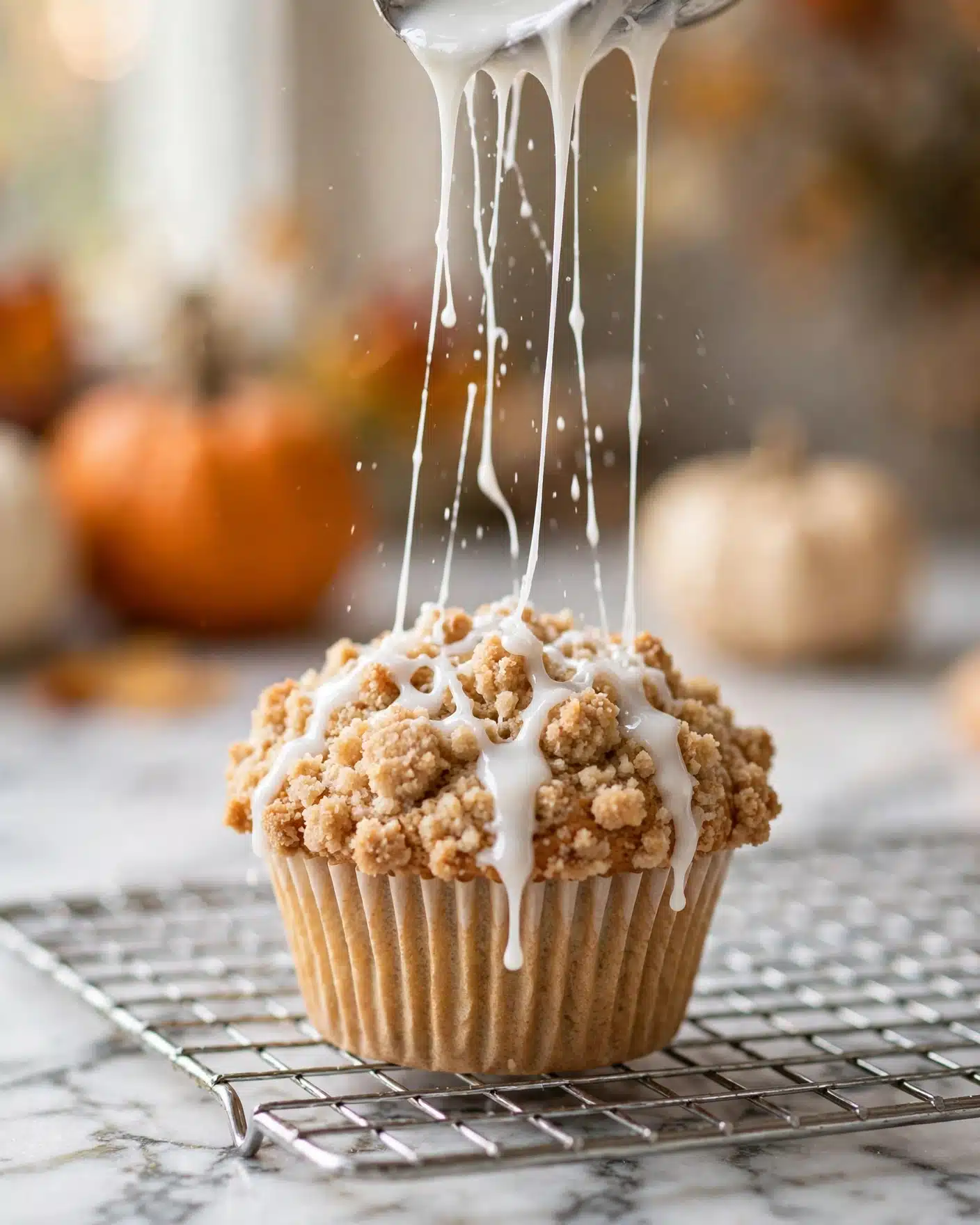 This image shows a close-up of a single crumb-topped muffin with a light brown crumb layer that looks soft and crumbly on top. White icing is being poured in thin streams over the crumbs from above, creating shiny, smooth lines that contrast with the crumbly texture beneath. The muffin is in a light beige paper cup with ridges, placed on a metal cooling rack which sits on a white marbled surface. In the blurry background, there are out-of-focus pumpkins adding an autumn feel to the scene. photo taken with an iphone --ar 4:5 --v 7