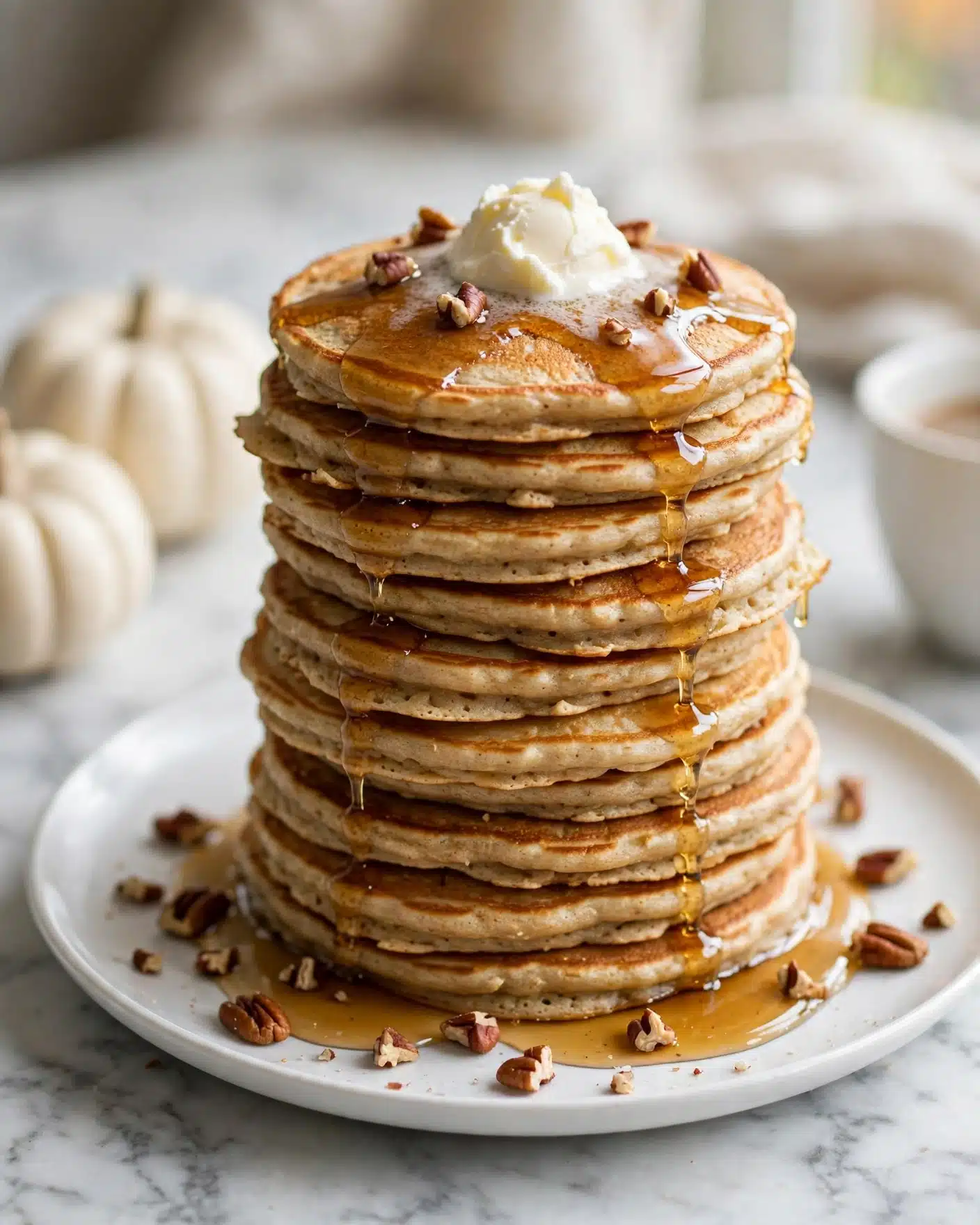 A tall stack of about ten light brown pancakes with a slightly uneven texture sits on a white plate, each pancake softly golden around the edges. On top, there is a dollop of creamy white butter melting slowly, drizzled with amber syrup that glistens as it runs down the sides. Small pieces of chopped pecans are scattered over the syrup and around the base of the pancakes. In the background, the scene is set on a white marbled surface with two small white pumpkins on the left and a white cup partially visible on the right. Photo taken with an iphone --ar 4:5 --v 7