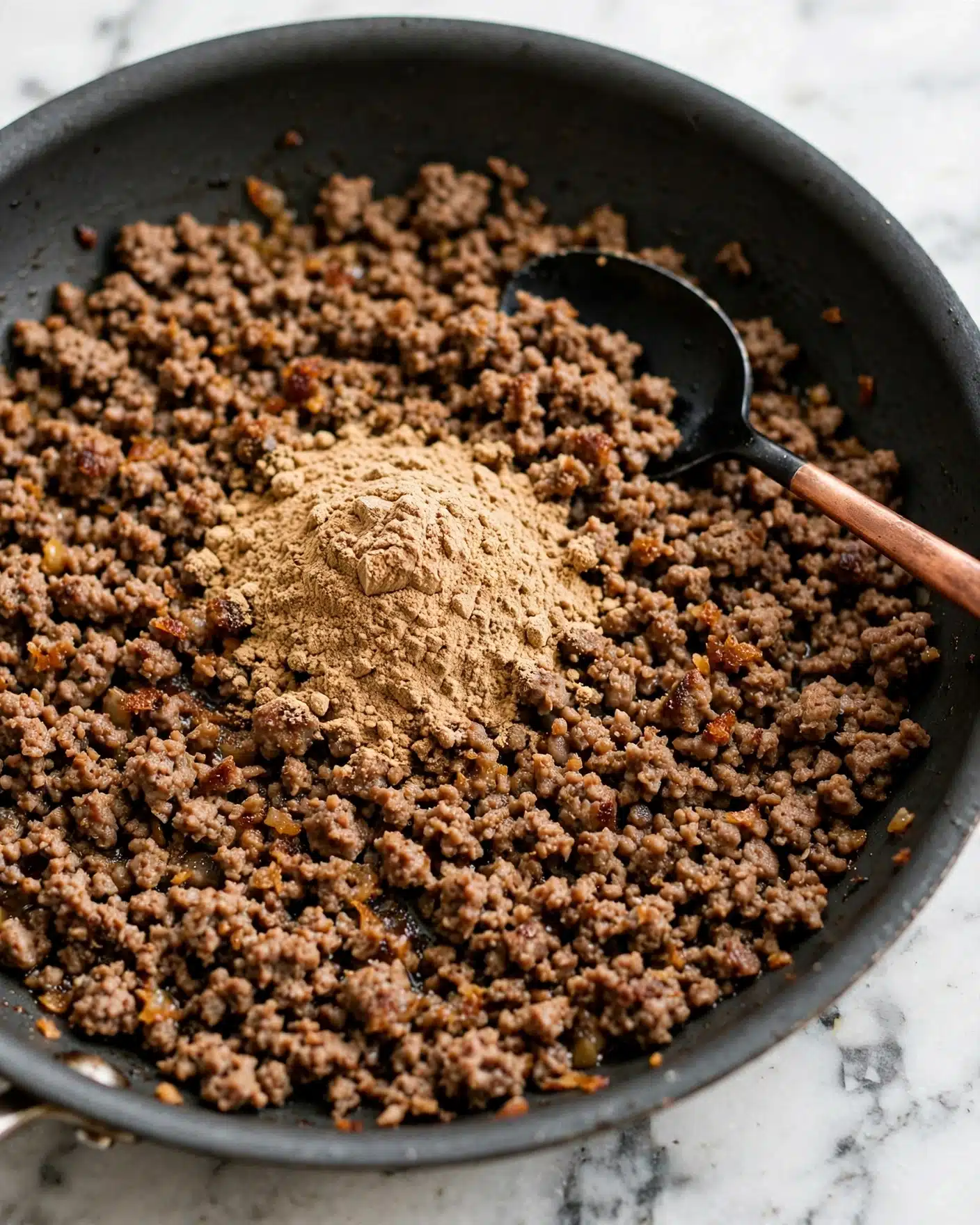 A close-up view of a dark gray frying pan filled with browned ground meat that has a crumbly texture, topped with a single mound of light brown powdered spice in the center. A black spoon with a copper handle rests inside the pan on top of the meat, slightly to the right side. The pan is placed on a white marbled surface. photo taken with an iphone --ar 4:5 --v 7