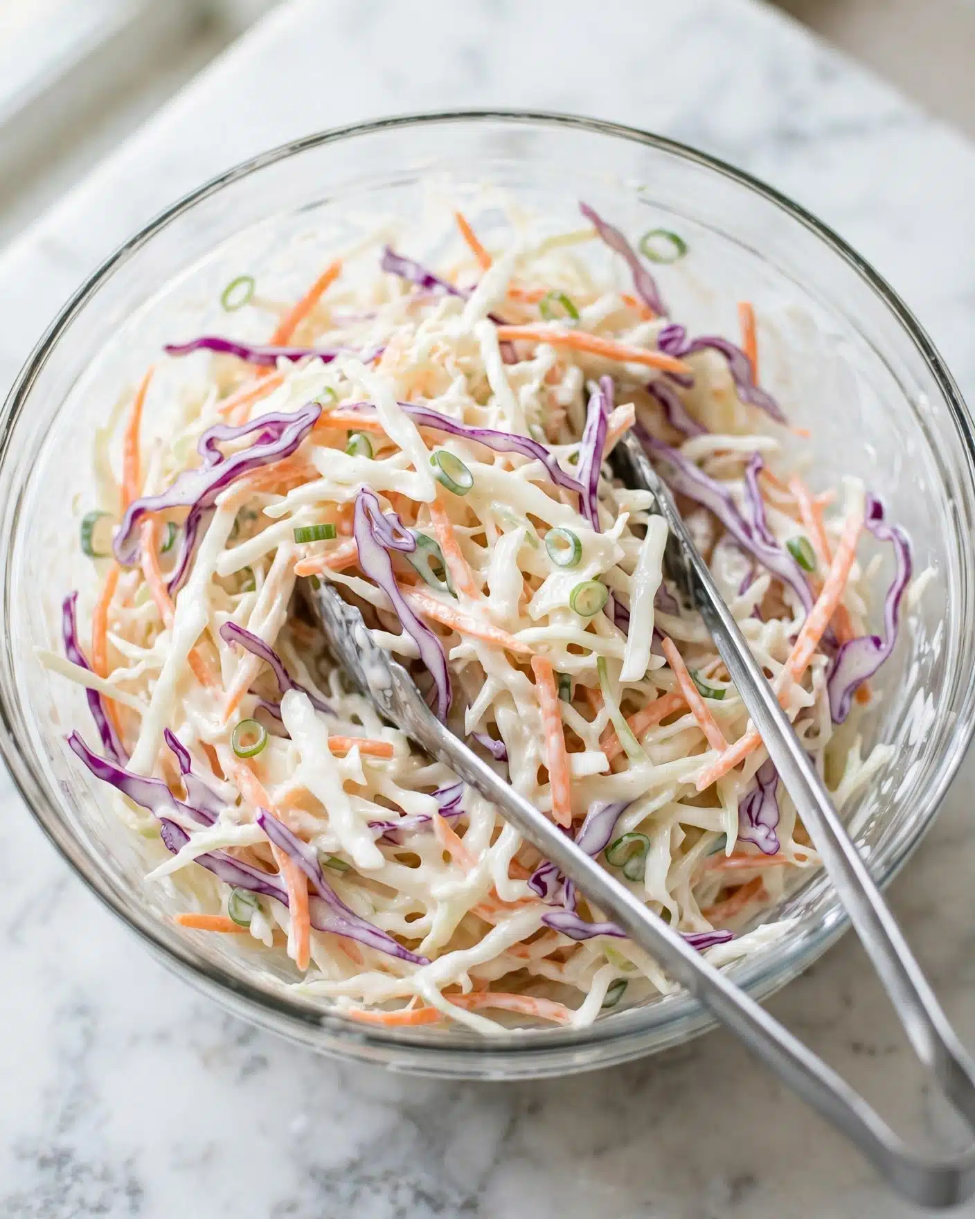 Classic Coleslaw Recipe 5 A clear glass bowl filled with a mix of shredded vegetables including white cabbage, light orange carrots, and light purple cabbage, all coated in a creamy white sauce. Thin green slices of scallion or similar greens are mixed in, adding dots of green throughout. The textures look soft and moist with a bit of creaminess from the sauce. Two long metal tongs rest inside the bowl, one angled towards the bottom right. The bowl sits on a white marbled surface, creating a clean and fresh look. photo taken with an iphone --ar 4:5 --v 7