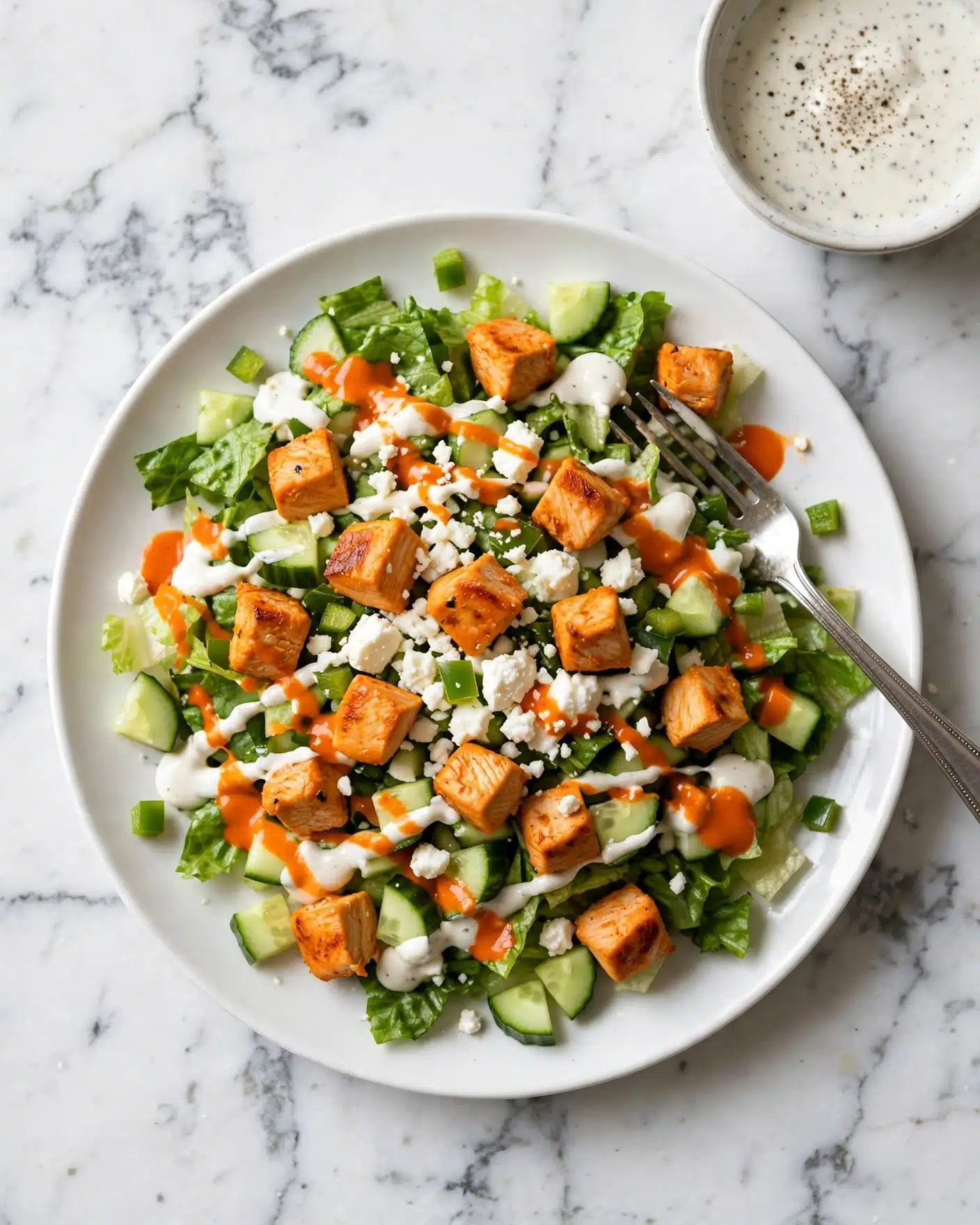A white plate holds a colorful mixed salad with layers of chopped green lettuce and cucumber forming the base, topped with evenly scattered orange cubes of grilled meat, crumbled white cheese, and small bits of green pepper. The salad is drizzled with white creamy dressing and bright orange sauce unevenly spread on top. A silver fork rests on the right side of the plate, partially inserted into the salad. The plate is placed on a white marbled surface with a bowl of white sauce speckled with black pepper in the top right corner. Photo taken with an iphone --ar 4:5 --v 7