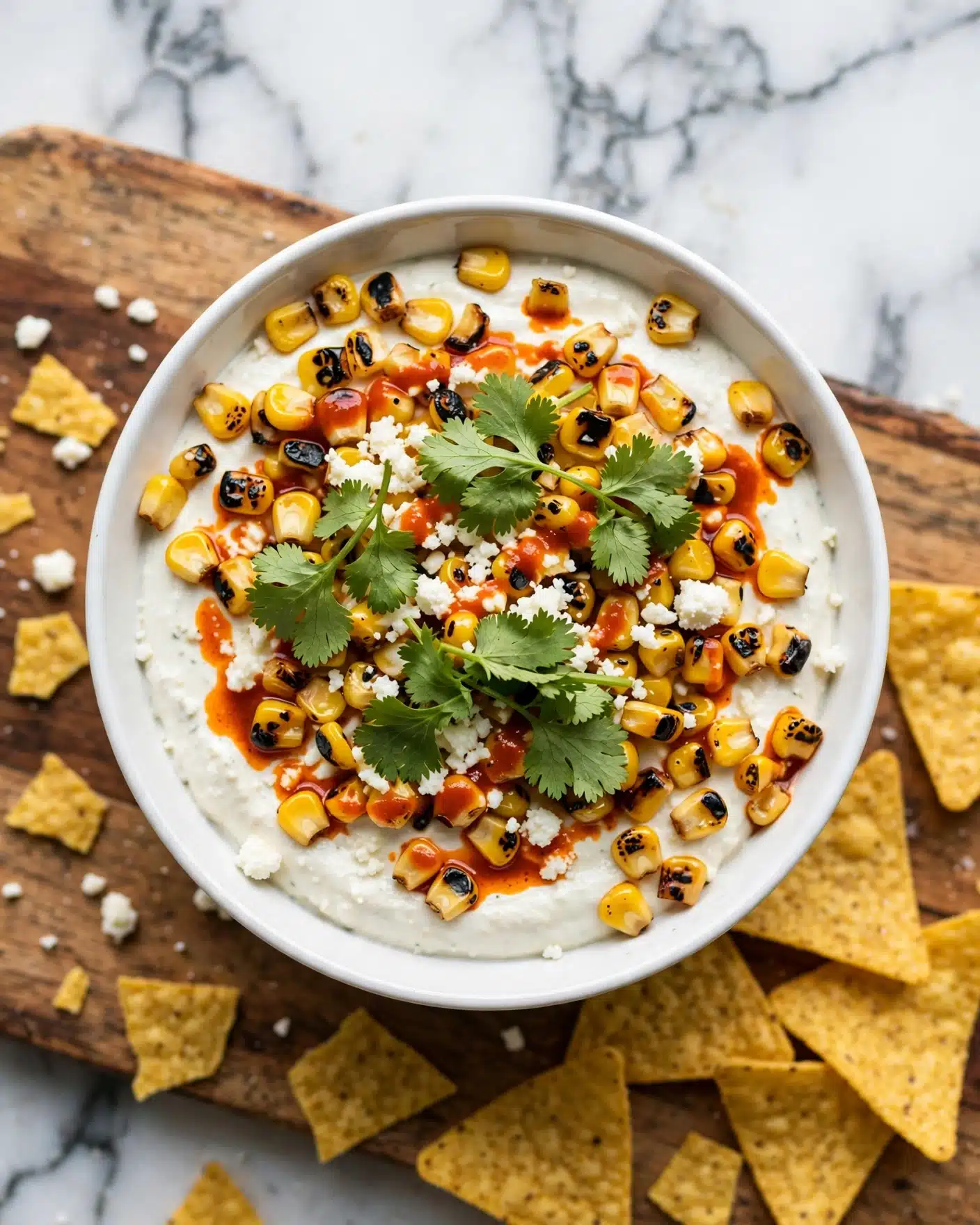 A white bowl filled with creamy white dip as the base layer, topped with grilled yellow corn pieces that have black char marks scattered evenly across the surface. The corn is mixed with a slight drizzle of orange-red oil or sauce, creating a contrast against the cream. Fresh green cilantro leaves are layered on top, along with small crumbles of white cheese sprinkled generously over the dish. The bowl is placed on a wooden board with scattered pieces of yellow tortilla chips around it. The background features a white marbled surface. photo taken with an iphone --ar 4:5 --v 7