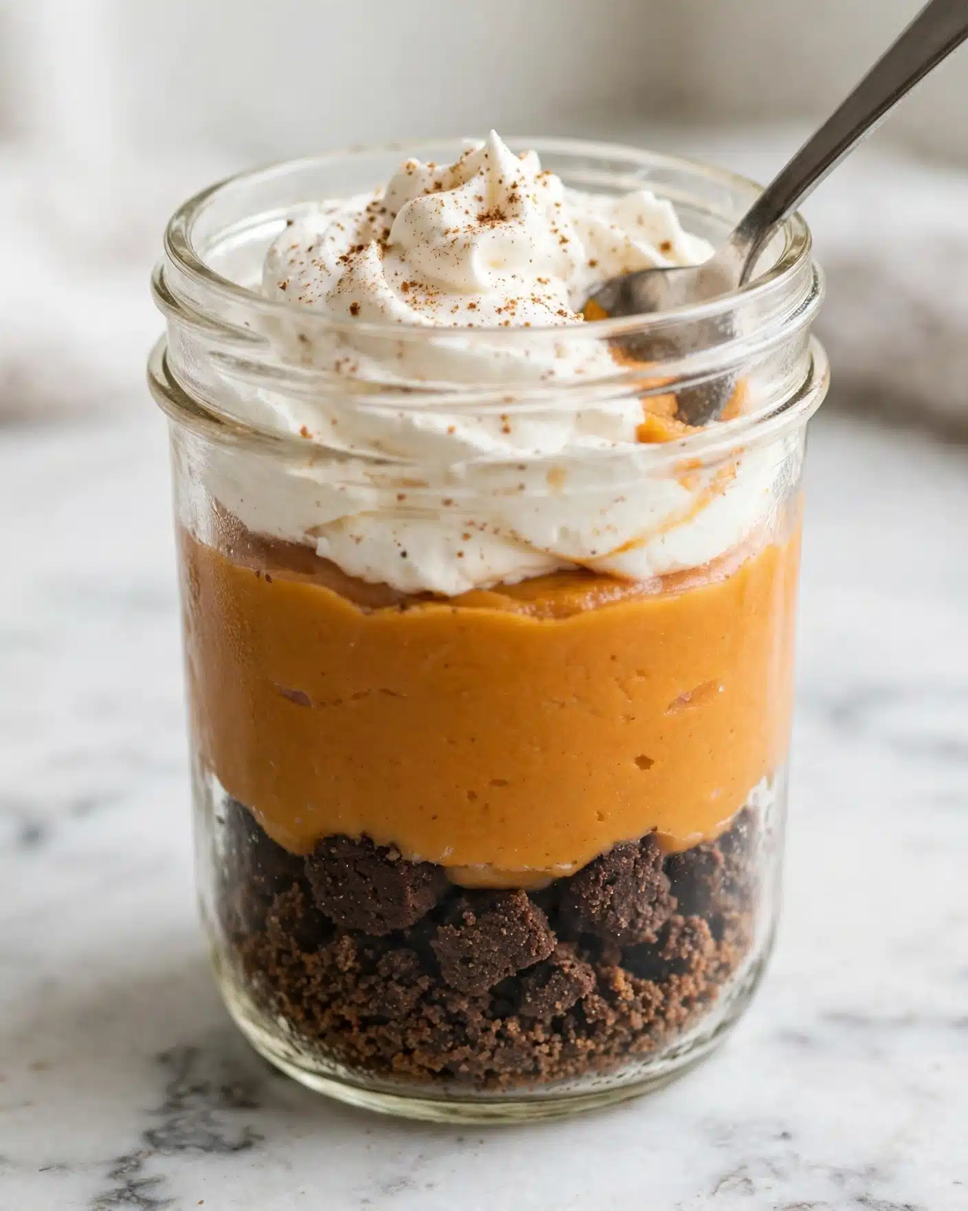 This image shows a clear glass jar with three visible layers. The bottom layer is a dark brown crumbly base, likely made of crushed cookies. The middle layer is thick and smooth, with a rich orange color resembling pumpkin or sweet potato filling. The top layer is white whipped cream, swirled with a smooth texture and sprinkled lightly with cinnamon or nutmeg. A silver spoon dips into the jar, pressing into the whipped cream and filling. The jar sits on a white marbled surface. Photo taken with an iphone --ar 4:5 --v 7
