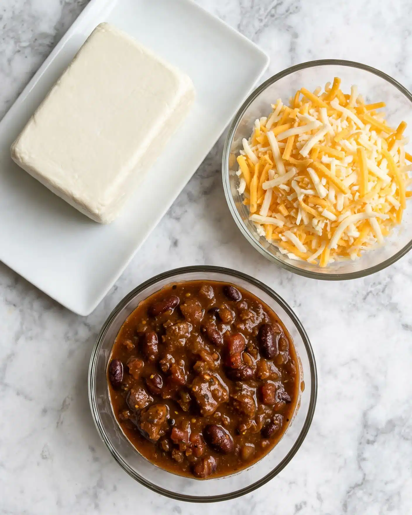 The image shows three bowls and a white rectangular plate on a white marbled surface. The top left white rectangular plate holds a large block of cream-colored soft cheese. To the right, there is a clear glass bowl filled with shredded cheese that is yellow and white mixed. Below, another clear glass bowl contains a thick brown chili mixture with visible beans and chunks, having a shiny, rich texture. The items are spaced apart and viewed from above. photo taken with an iphone --ar 4:5 --v 7