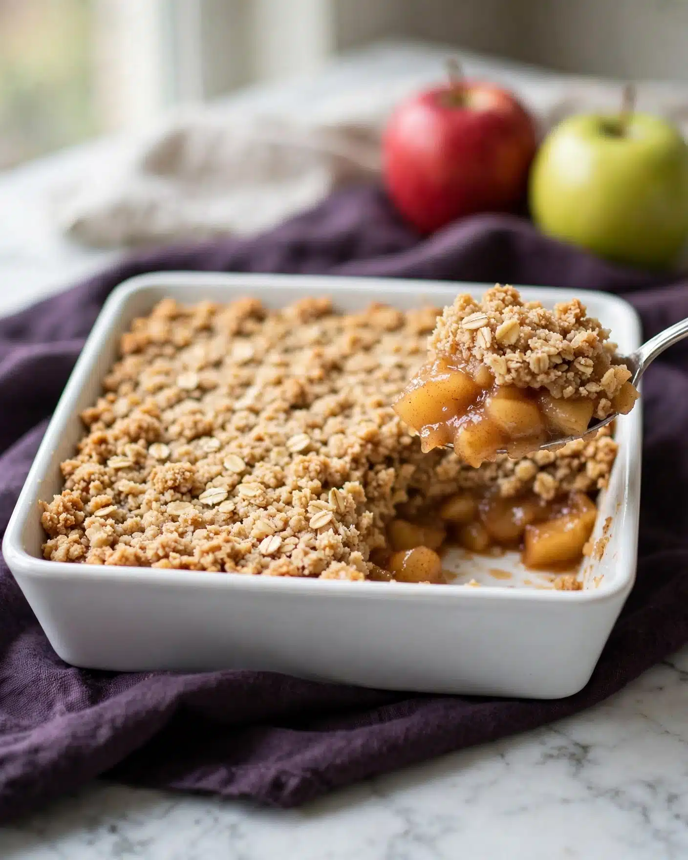 A white square baking dish holds a two-layer apple crisp dessert. The bottom layer is soft, cooked apple pieces with a warm golden color and a hint of cinnamon, visible at the lifted side where a silver spoon is scooping a portion. The top layer is a thick, slightly crumbly oat topping with a light golden-brown color and texture made of oats and small bread crumbs. The dish rests on a dark purple cloth over a white marbled surface. In the soft background, two whole apples, one red and one green, add a pop of color. photo taken with an iphone --ar 4:5 --v 7