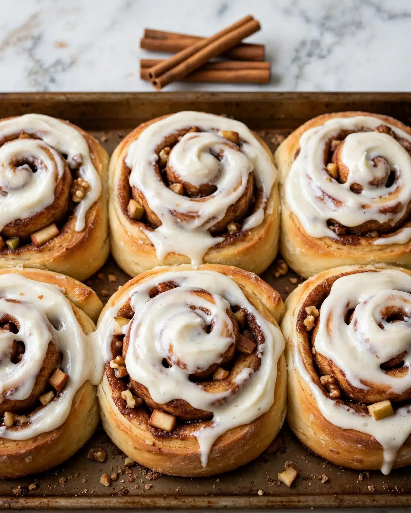 A close-up view of six cinnamon rolls arranged closely on a brown baking tray placed on a white marbled surface. Each roll has a thick, creamy white frosting layer spread on top, with visible swirls of golden-brown dough rolled around a cinnamon sugar filling that shows a rich dark brown color inside. Small chunks of nuts or apple pieces are scattered within the cinnamon layers, adding texture. In the background, three cinnamon sticks are stacked casually near the top edge of the tray. Photo taken with an iphone --ar 4:5 --v 7