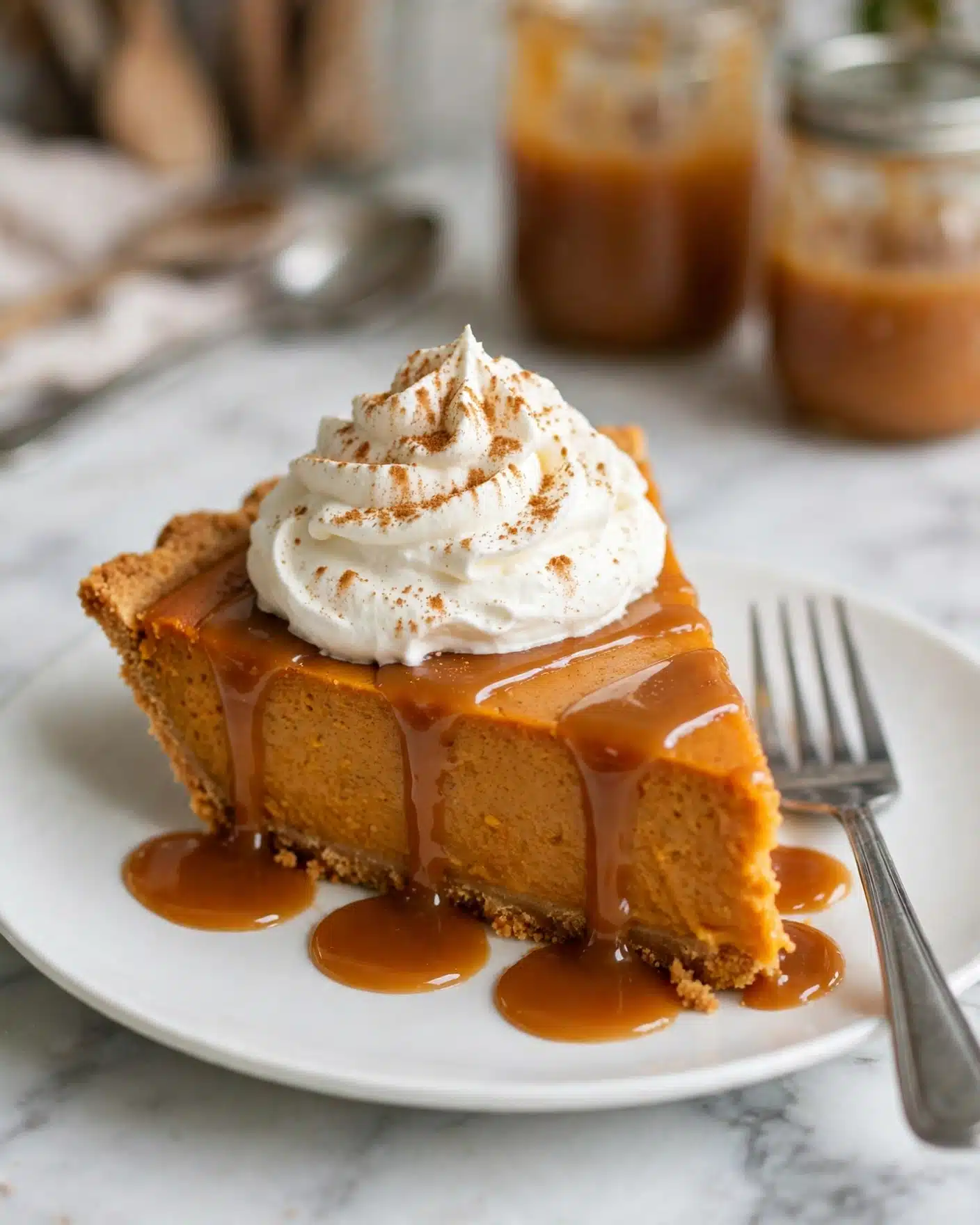 A single slice of orange pumpkin pie sits on a white plate with a graham cracker crust visible at the base. The slice is topped with a thick swirl of white whipped cream sprinkled with light brown cinnamon powder. A rich, glossy caramel sauce flows down the sides of the pie, pooling around the bottom on the plate. A silver fork rests beside the slice on the right side of the plate. The background features a soft white marbled texture with a blurred glass jar of caramel sauce and utensils in the distance. Photo taken with an iphone --ar 4:5 --v 7