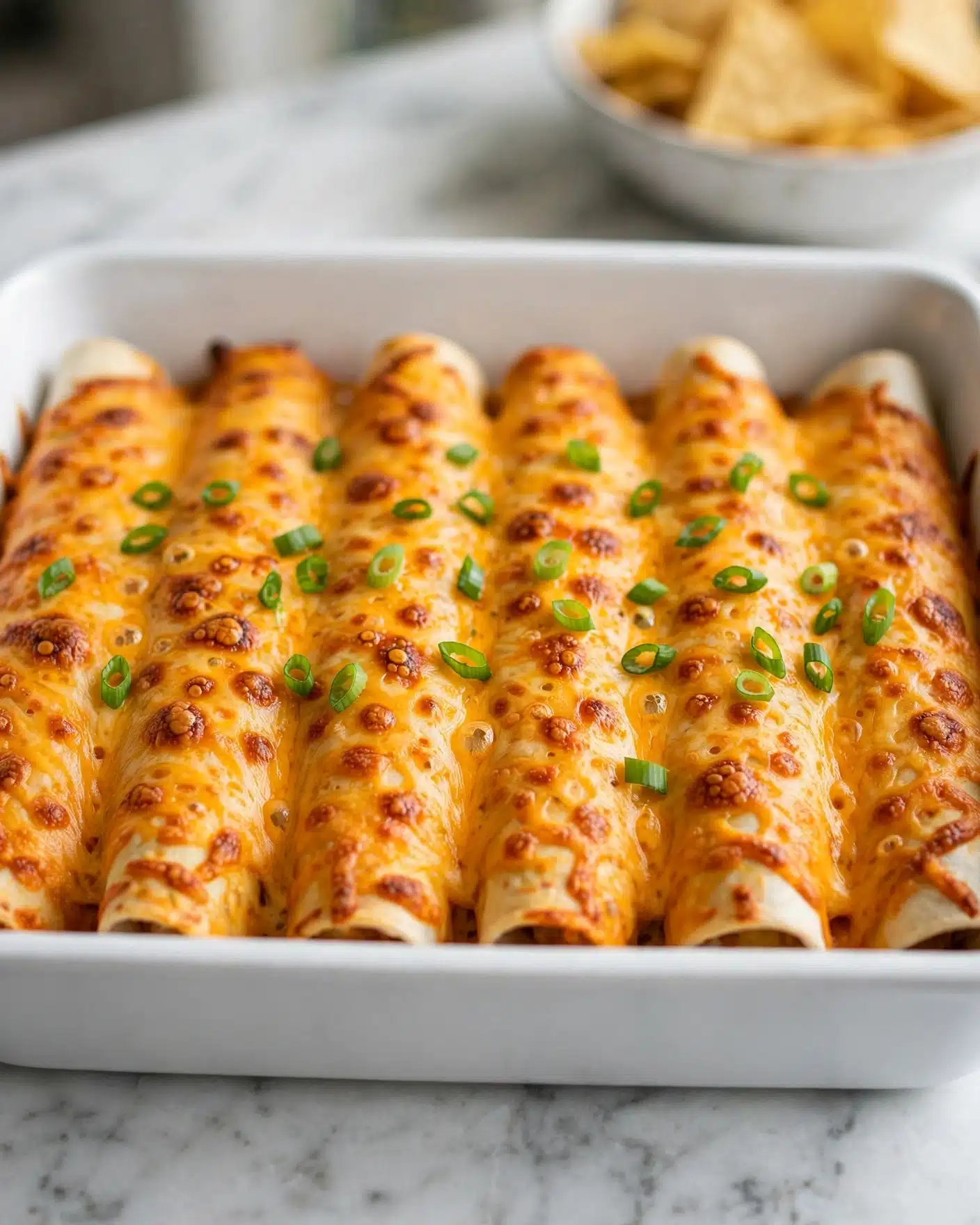 A white rectangular baking dish holds six rolled enchiladas arranged side by side, each covered with a layer of melted golden orange cheese with some browned spots. The cheese layer has a slightly bubbly texture and covers the enchiladas fully. Bright green sliced scallions are sprinkled evenly over the cheese as a garnish. The dish is placed on a white marbled surface with part of a blurred white bowl of tortilla chips visible in the background. photo taken with an iphone --ar 4:5 --v 7