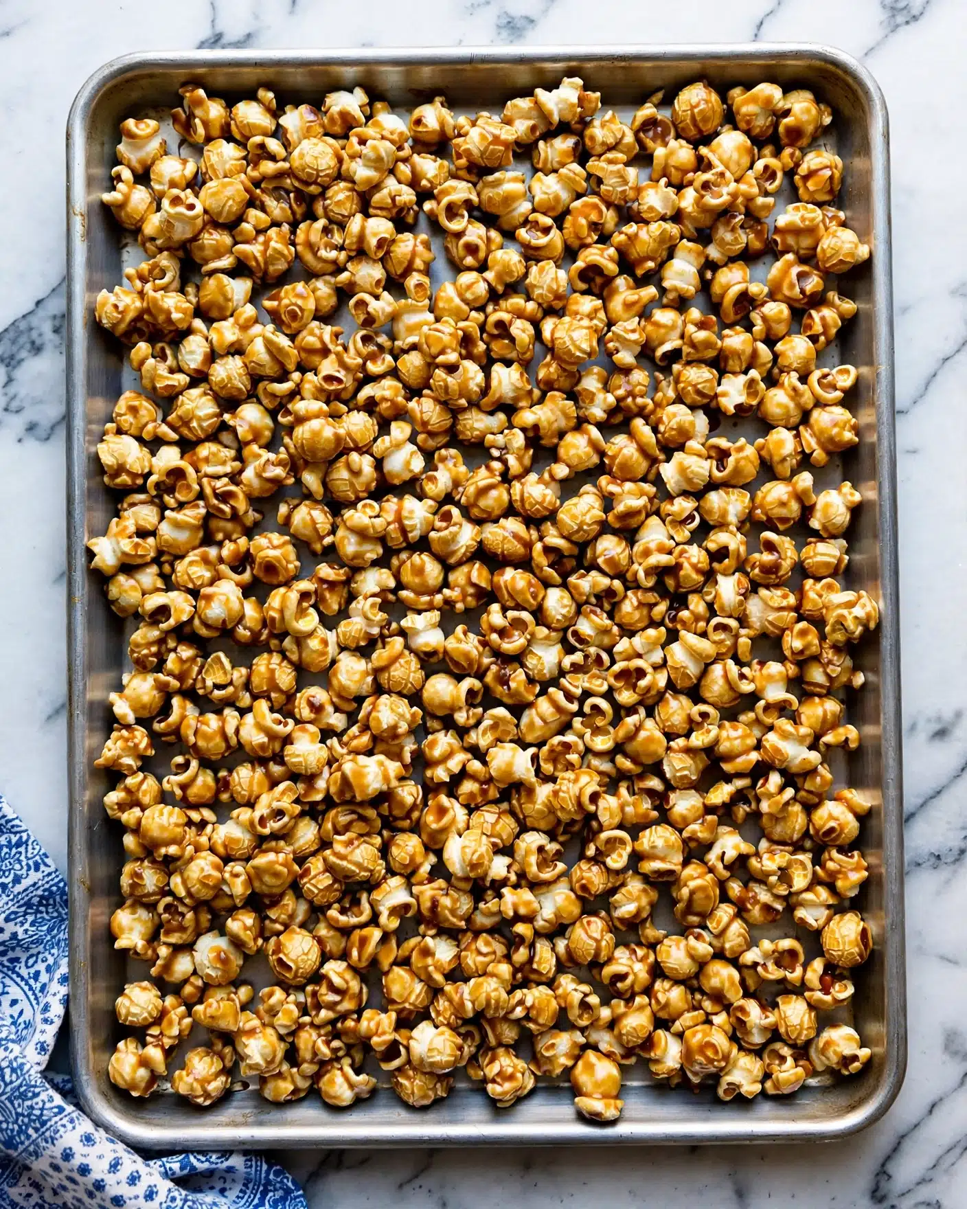 A large metal baking tray filled with a single thick layer of caramel popcorn, where each piece is coated in a shiny, golden-brown caramel glaze with a mix of lighter and darker spots, showing a slightly bumpy texture. The tray is placed on a white marbled surface, and a corner of a blue and white patterned cloth is visible at the bottom left. The popcorn pieces are irregular in shape, tightly packed but not overlapping much, creating a rough but even spread across the tray. photo taken with an iphone --ar 4:5 --v 7