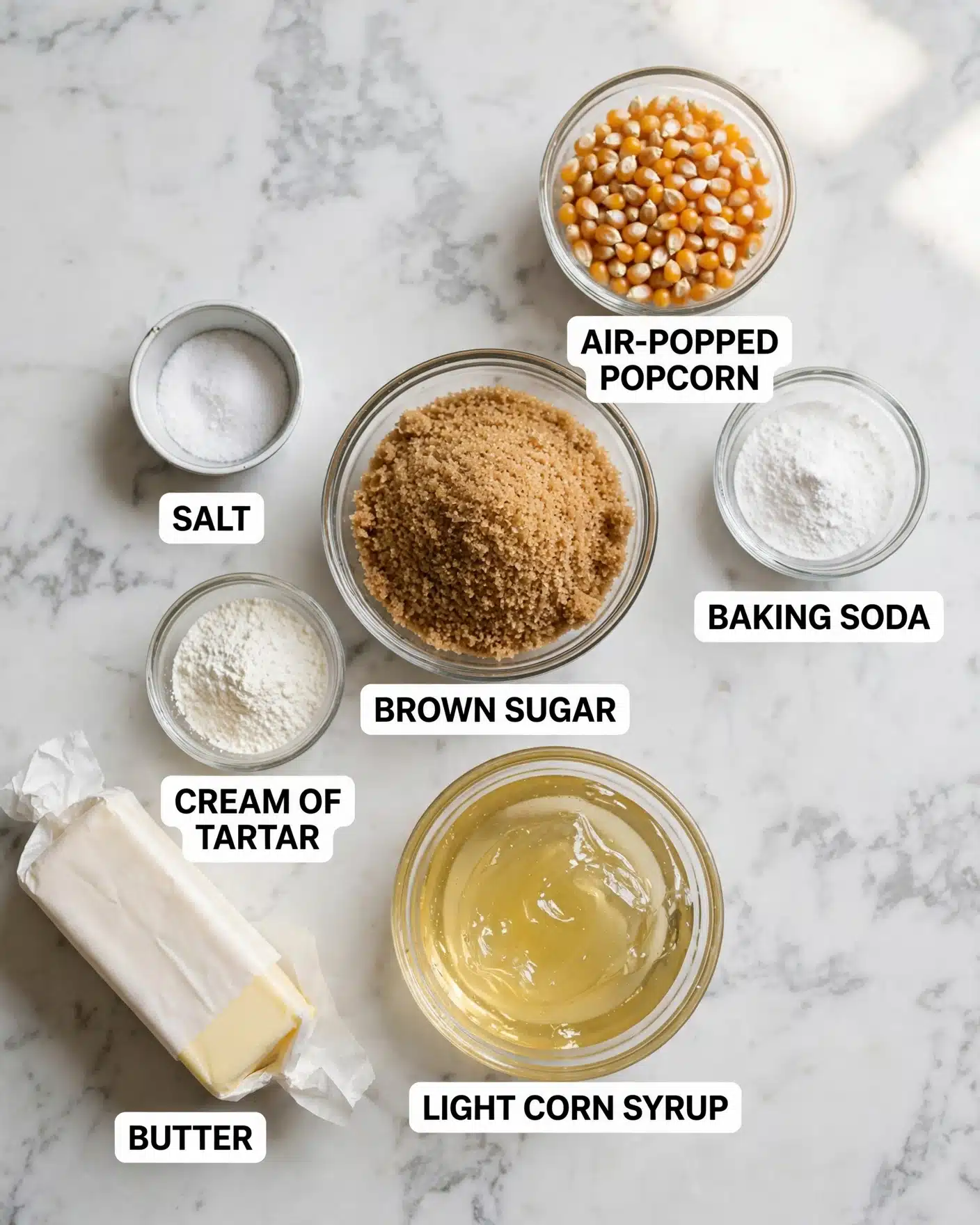 The image shows six ingredients on a white marbled surface arranged in a loose circle. At the center is a clear glass bowl filled with a large amount of light brown sugar granules with a rough texture. Above it and slightly to the right, there is a clear glass bowl holding yellow popcorn kernels with a smooth, shiny surface. Near the popcorn is a small clear glass bowl containing fine white baking soda powder. To the left of the brown sugar bowl, there is a small white metal bowl with fine white salt. Below the salt, a small clear glass bowl holds white cream of tartar powder. Below the brown sugar bowl is a clear glass bowl with a thick, clear, light corn syrup. On the bottom left, there is a stick of butter, pale yellow in color, partially wrapped in white wax paper. Each ingredient is labeled with bold black text on a white block: 