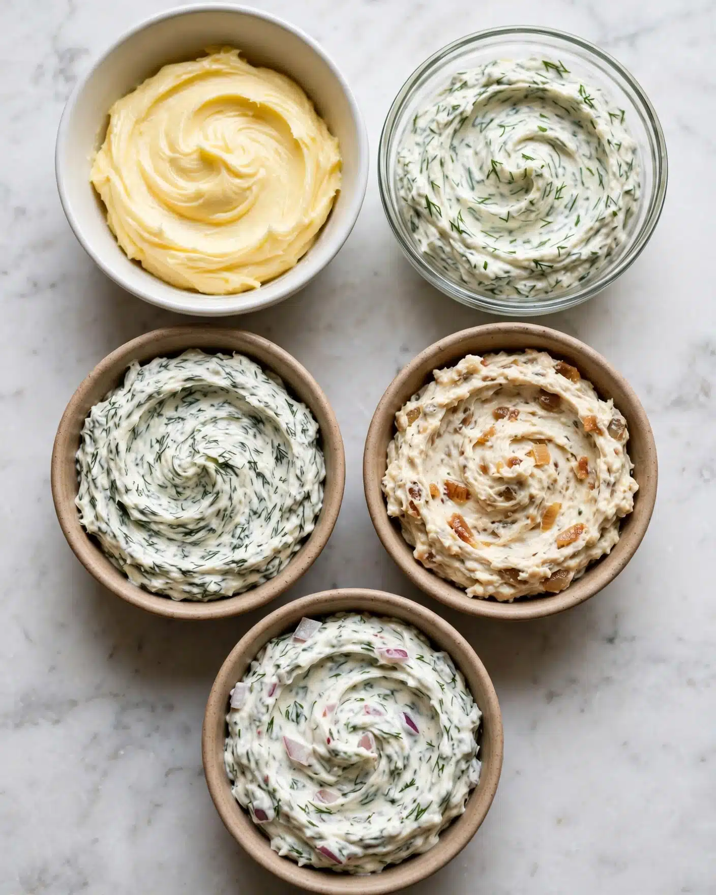 Five small bowls filled with different kinds of soft spreads or butters are placed on a white marbled surface. The top left bowl is white and contains smooth pale yellow butter with some textured swirling on top. Next to it on the right is a clear glass bowl filled with a creamy white spread mixed with green herbs, showing a soft texture with swirls. Below these, two small light brown ceramic bowls sit side by side; the left one has a white spread mixed with dark green herbs with swirling patterns, while the right one holds a light cream spread with small bits of caramelized onions mixed in, showing a rougher texture. At the bottom, there is another light brown ceramic bowl with white spread mixed with both green herbs and small pieces of reddish onion, also swirled softly on top. photo taken with an iphone --ar 4:5 --v 7