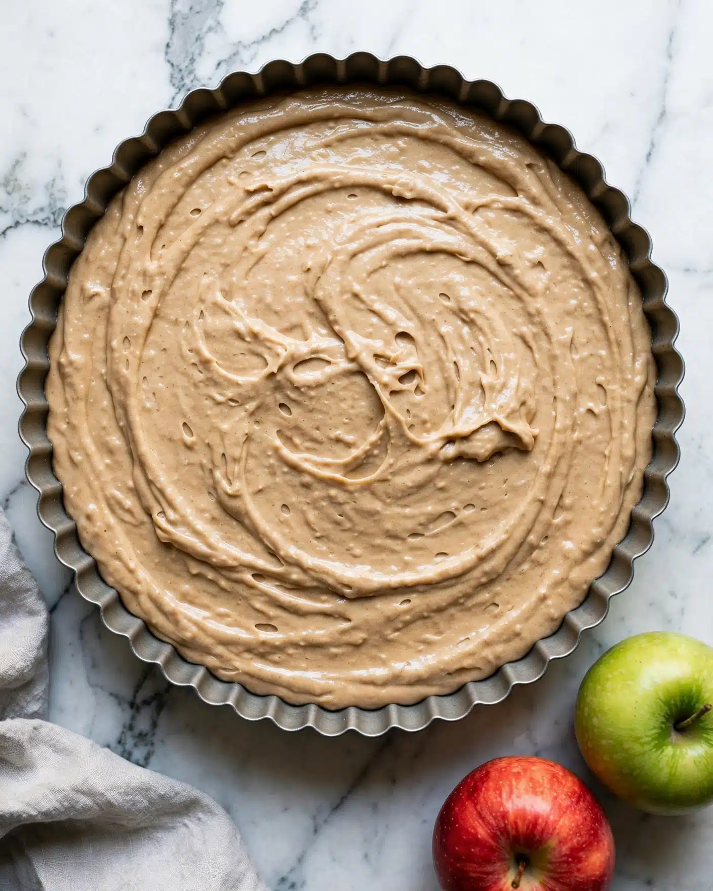 A round baking pan with scalloped edges is filled with a smooth light brown batter that has a creamy and slightly thick texture. The batter spreads evenly inside the pan, showing slight swirls and gentle peaks. The pan sits on a white marbled surface with two apples, one red and one green, placed near the bottom right corner, and a light grey cloth partially visible on the bottom left. Photo taken with an iphone --ar 4:5 --v 7