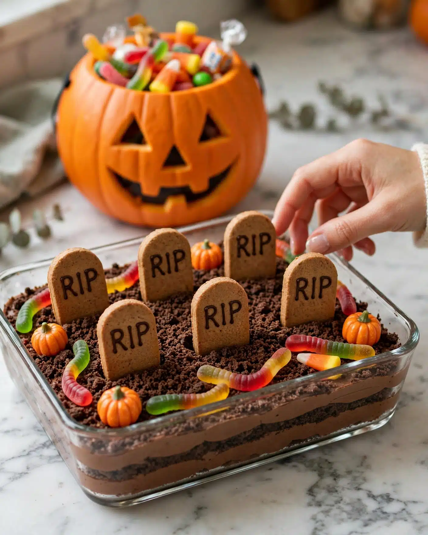 A clear glass rectangular dish filled with layered chocolate dessert topped with crumbled cookie crumbs resembling soil. Six oval cookie pieces with 