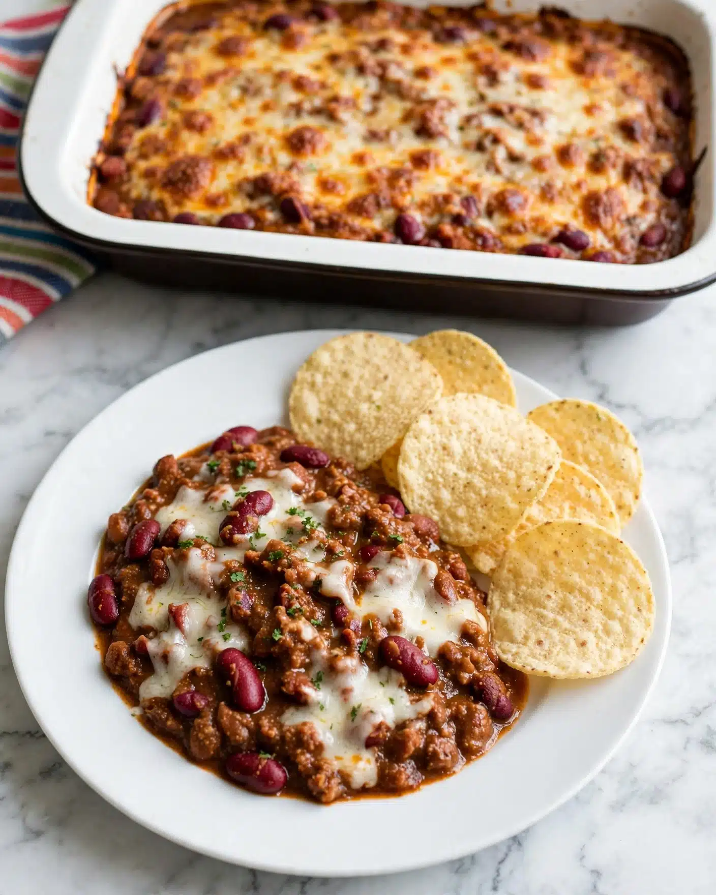 On a white plate set on a white marbled surface, there is a serving of chili that has a thick, rich brown sauce with visible red kidney beans and melted white cheese mixed in. Next to the chili, several light yellow, round tortilla chips with a dry, slightly rough texture are arranged. Behind the plate, there is a white baking dish with a dark outer rim filled with baked chili topped with melted, golden-brown cheese. Part of a striped cloth napkin with red, blue, green, and orange lines is visible near the baking dish. Photo taken with an iphone --ar 4:5 --v 7