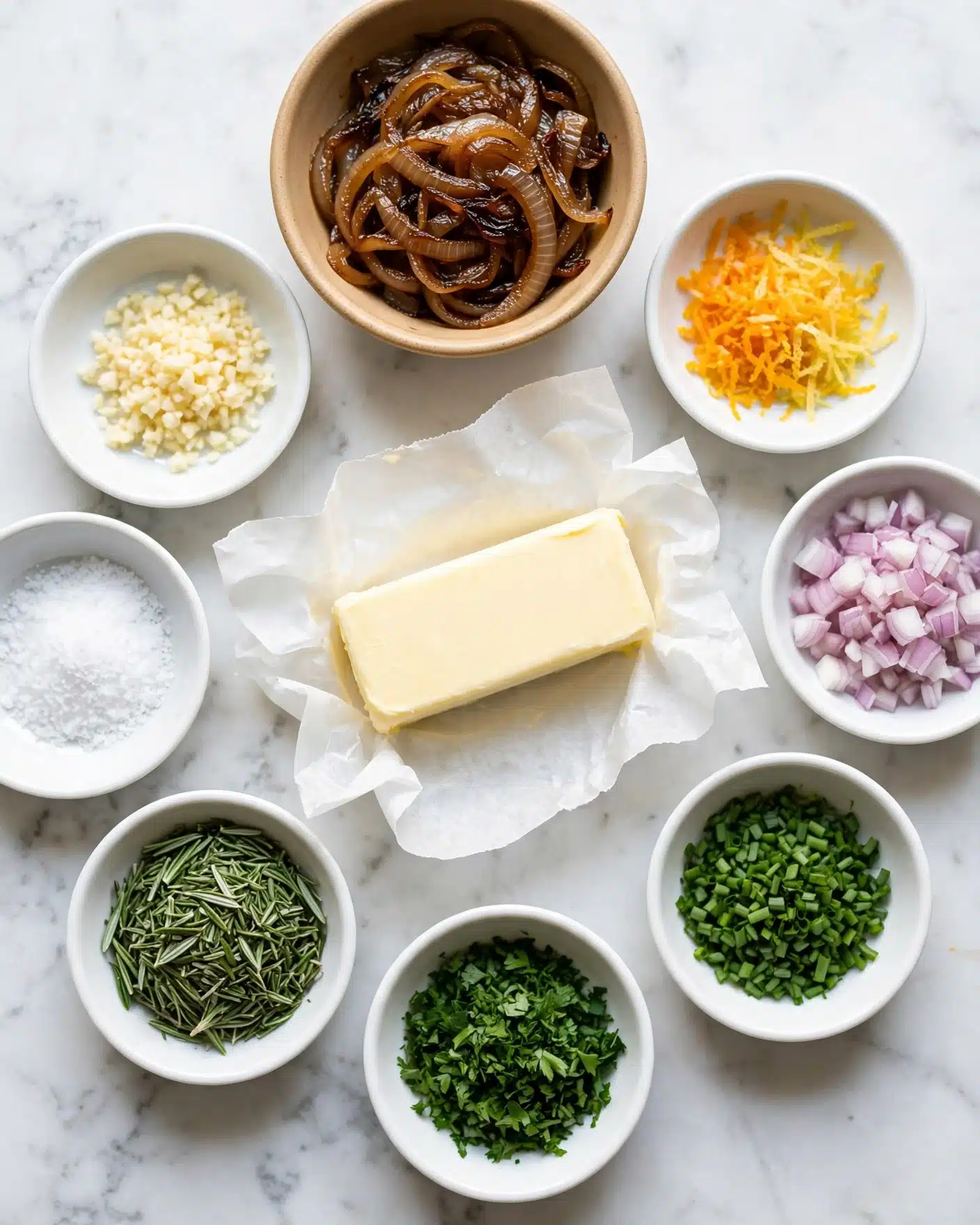 The image shows an arrangement of cooking ingredients on a white marbled surface. In the center is a stick of pale yellow butter wrapped in white parchment paper. Around it are eight small white bowls and one tan bowl, each containing different ingredients: finely chopped garlic, caramelized brown onions, white salt, chopped green rosemary, chopped green parsley, finely chopped green chives, diced light purple shallots, and a bowl with both bright orange and pale yellow citrus zest. The colors range from white, green, brown to orange, all neatly placed in bowls forming a loose circle around the butter. The lighting is bright and natural, giving a fresh and clean feel. Photo taken with an iphone --ar 4:5 --v 7