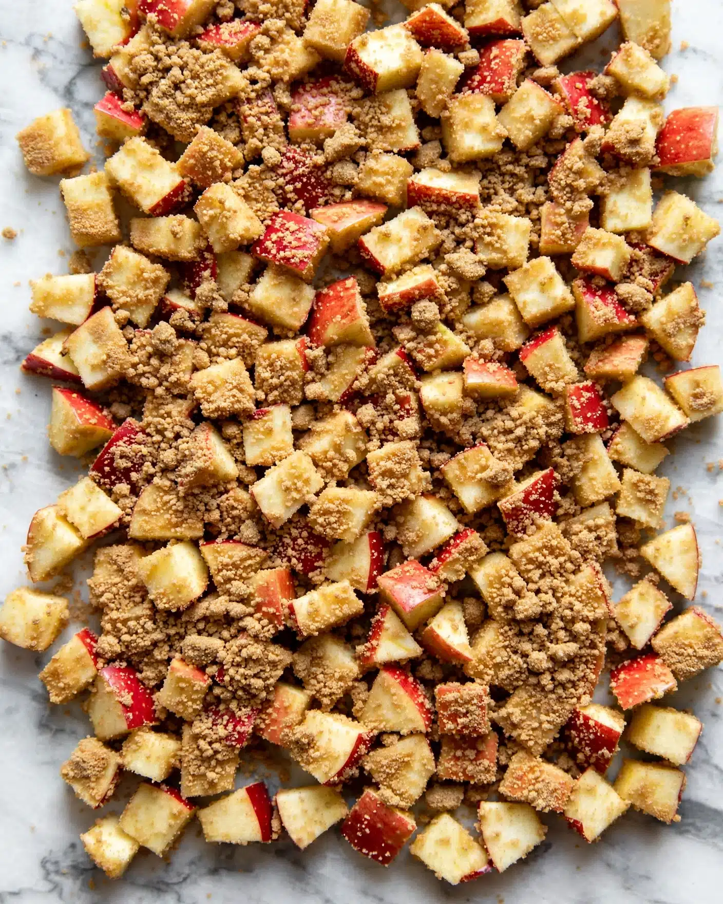 The image shows small, light yellow apple pieces with red skins scattered and covered evenly by a layer of fine brown sugar mixture. The texture is granular and slightly clumpy, covering most of the apple pieces but still letting the color of the apples show through. The apples are cut into small cubes and are spread closely together, forming one textured layer on a white marbled surface. photo taken with an iphone --ar 4:5 --v 7