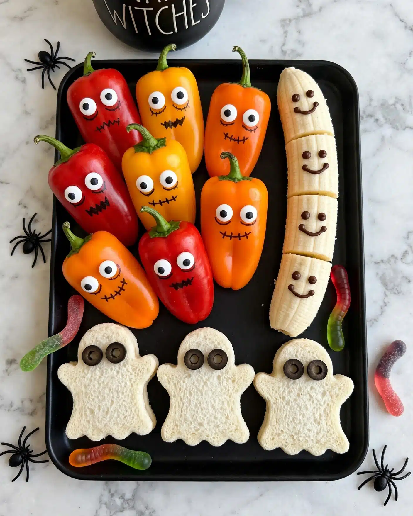 Halloween Kid-Friendly Snack Board Recipe 5 A black serving tray on a white marbled surface holds colorful mini bell peppers with large candy eyes, arranged in a cluster showing red, yellow, and orange colors with green stems on top. To the right are peeled bananas decorated with small chocolate faces, positioned in a vertical row. At the bottom, ghost-shaped white sandwich bread pieces sit upright, each with two black olive slices for eyes. Small black plastic spiders and candy worms add decoration around the tray edges. A black mug with white text