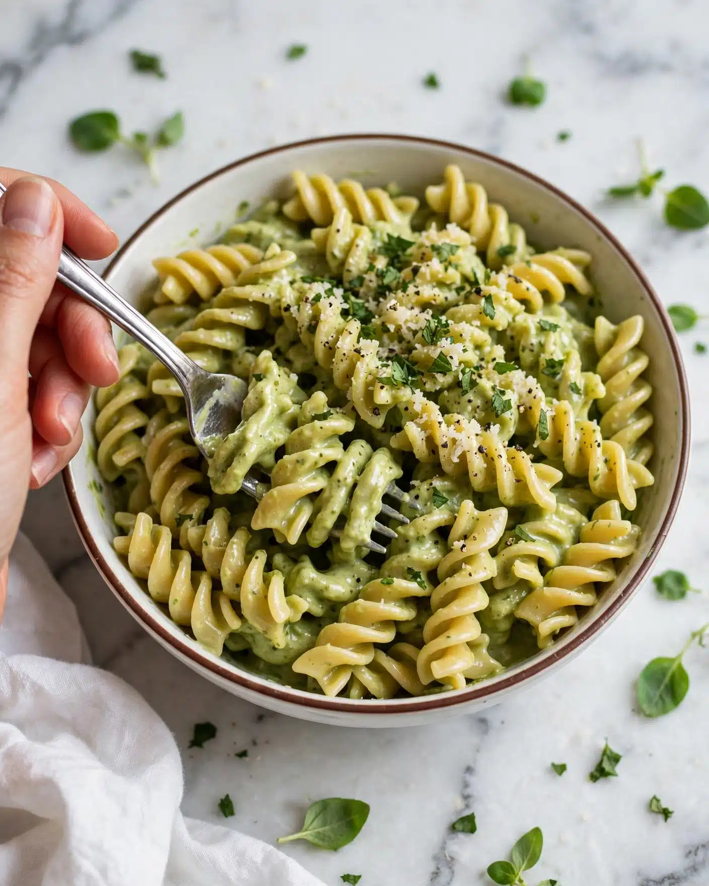 A close-up view of a bowl filled with curly, yellow pasta coated in a creamy light green sauce, garnished with finely chopped fresh green herbs and a sprinkle of grated cheese and black pepper on top. The pasta sits in a white bowl with a thin brown rim, placed on a white marbled surface. A silver fork is partly buried in the pasta with a woman's hand holding it on the left side. Some green leaves are scattered around the bowl, and a white cloth is partially visible near the bottom left corner. photo taken with an iphone --ar 4:5 --v 7