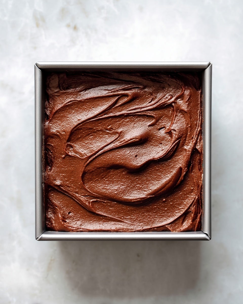 A single square layer of smooth, dark brown chocolate batter fills a silver square baking pan. The surface has thick, soft swirls and a slightly shiny texture, showing it is freshly spread and ready to be baked. The pan rests on a white marbled textured surface with a soft natural light coming from the side. photo taken with an iphone --ar 4:5 --v 7