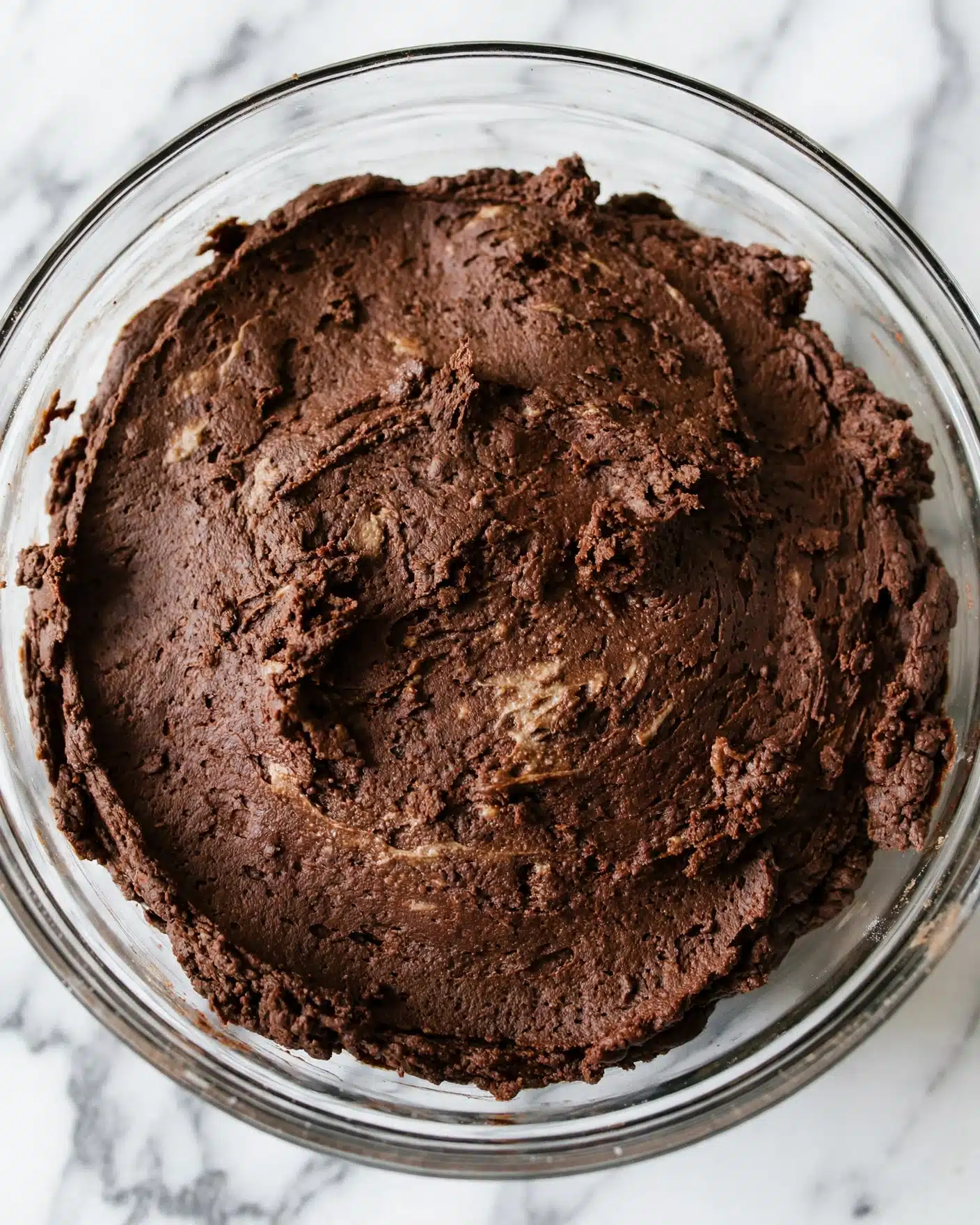 A close up top view of a clear glass bowl filled with a single thick layer of dark brown, smooth and dense chocolate dough, with some rough and creamy texture on the surface. The bowl sits on a white marbled texture. photo taken with an iphone --ar 4:5 --v 7