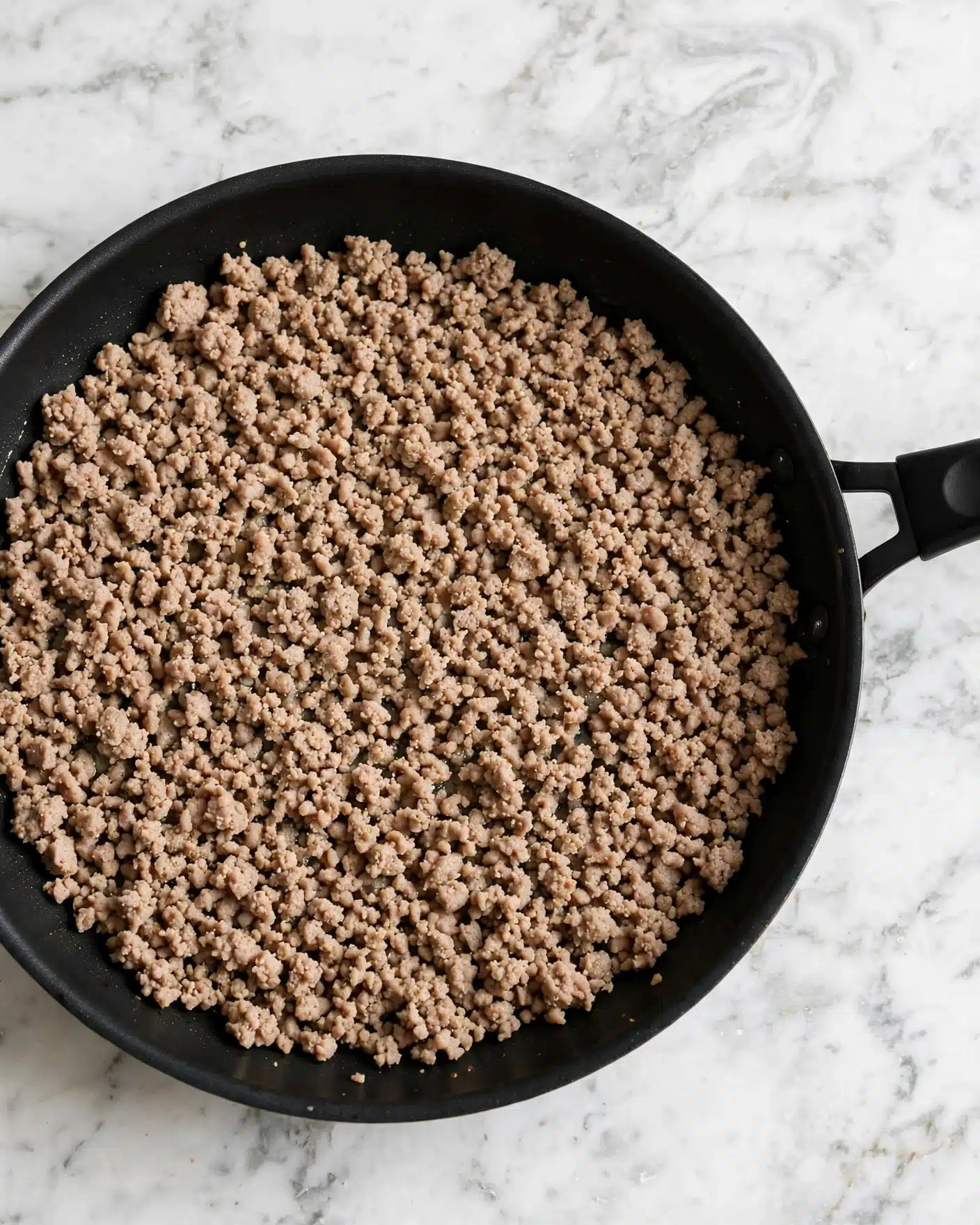 The image shows a black pan filled with light brown cooked ground meat that looks soft and crumbly. The meat covers the entire pan surface evenly with small bits and texture visible. The handle of the pan is black and positioned on the right side. The pan is placed on a white marbled surface. photo taken with an iphone --ar 4:5 --v 7